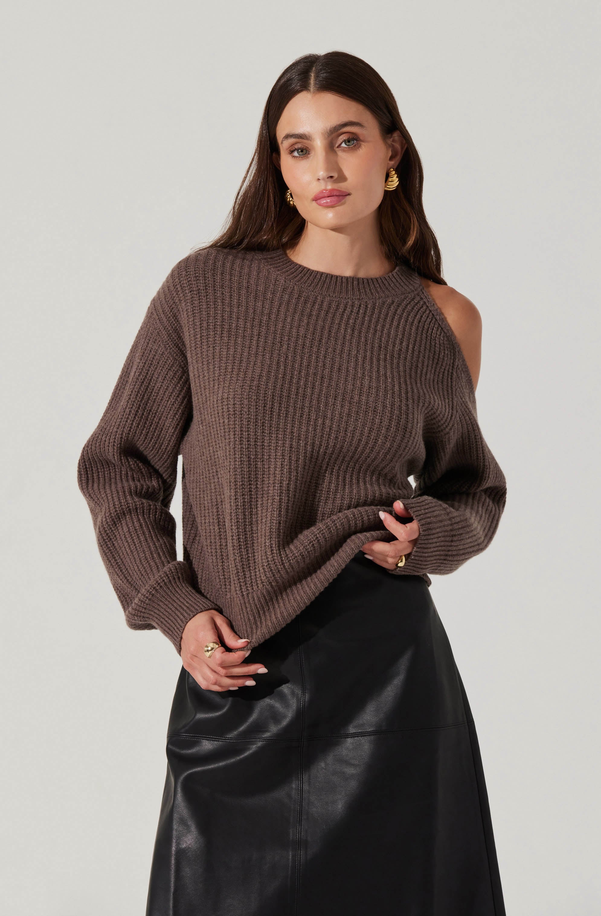 A woman is wearing a loose, oversized, brown knit sweater with a ribbed pattern, featuring a boat neckline that exposes her left shoulder. She has paired it with a high-waisted, black leather skirt. She accessorizes with gold hoop earrings and rings on her fingers.