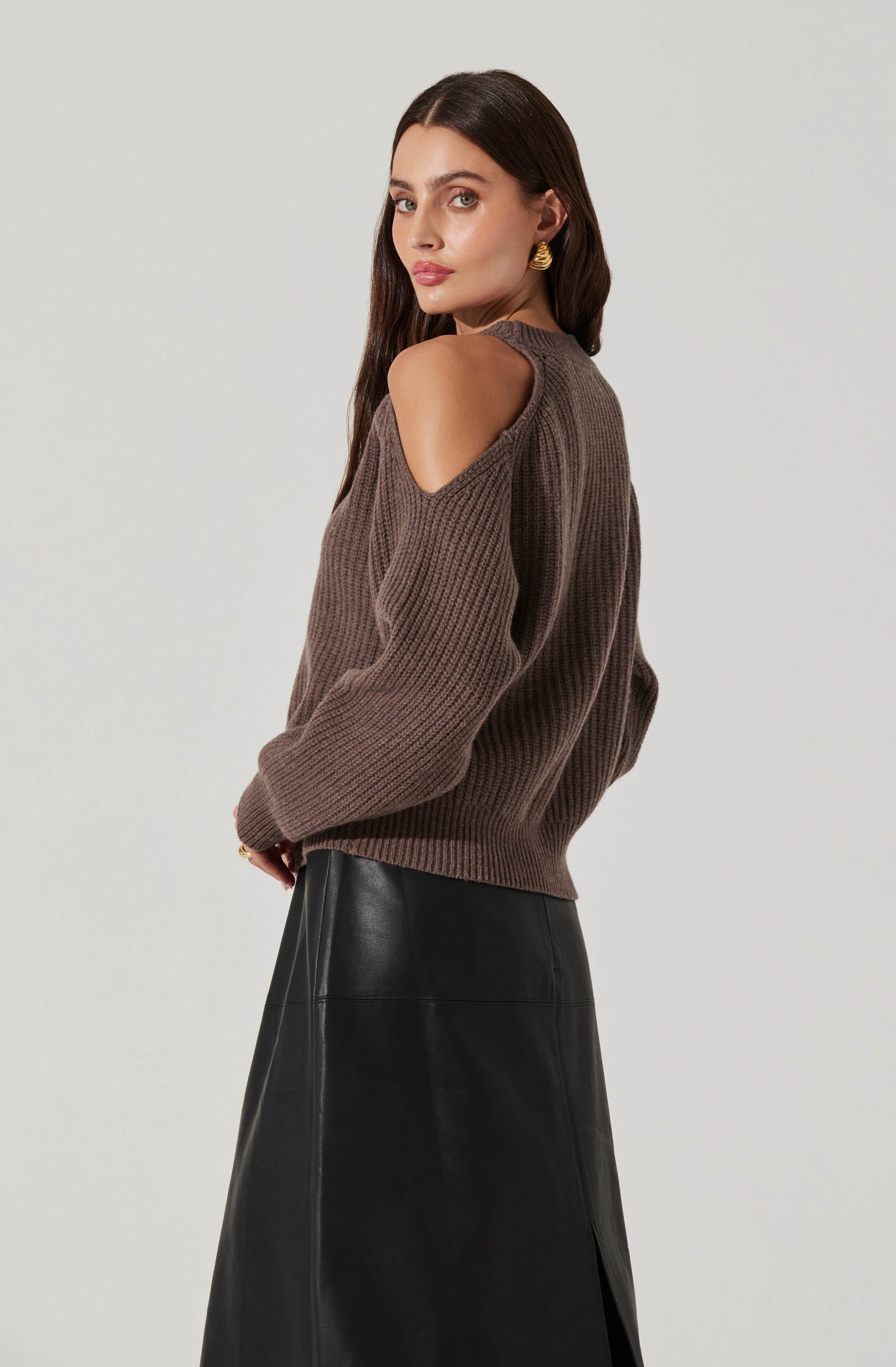A woman is wearing a brown knitted sweater with shoulder cut-outs and long sleeves, paired with a long black leather skirt. She has long, straight dark hair and is accessorized with large gold earrings.