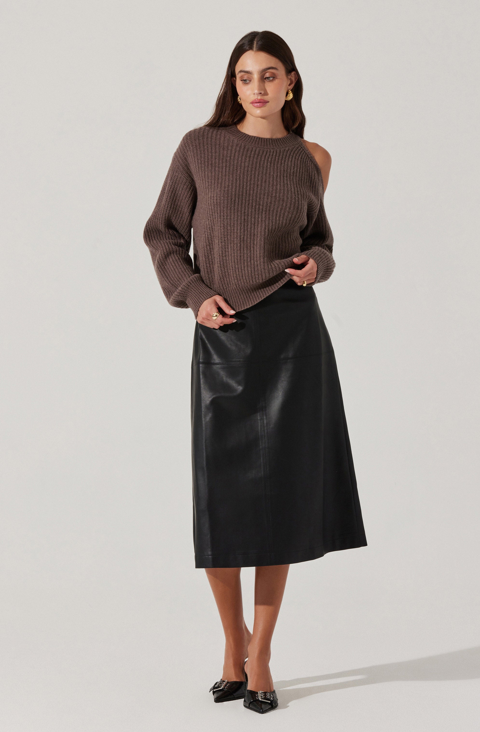 A woman is wearing a taupe, long-sleeved, ribbed knit sweater with a slight off-shoulder design on one side. She pairs it with a black, high-waisted, midi-length leather skirt that has a smooth, shiny finish. She completes the look with black pointed-toe shoes featuring embellishments on the front.
