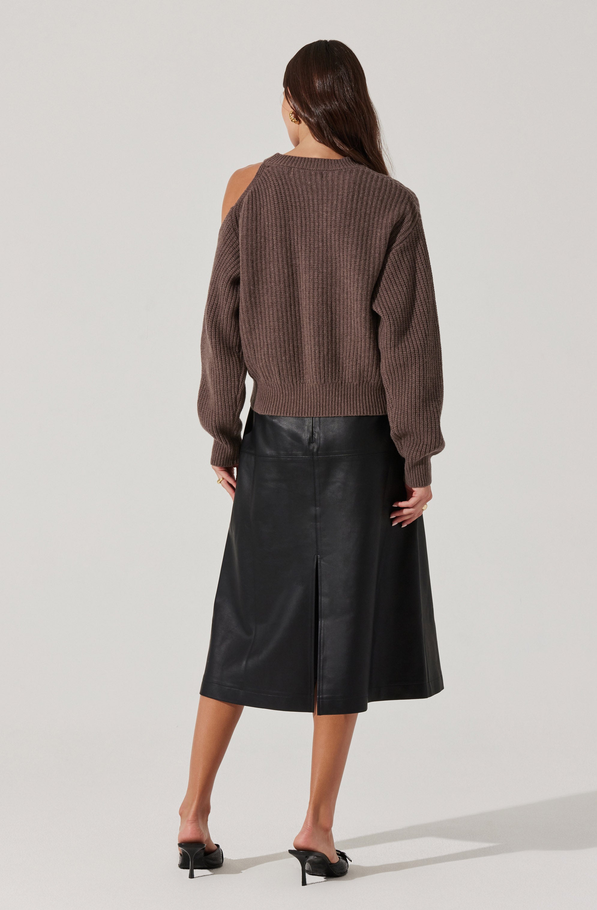 A woman is wearing an oversized, chunky knit sweater in a brown color, with long sleeves and a crew neckline. She pairs it with a high-waisted, black leather midi skirt with a front slit, and black open-toe heeled mules.