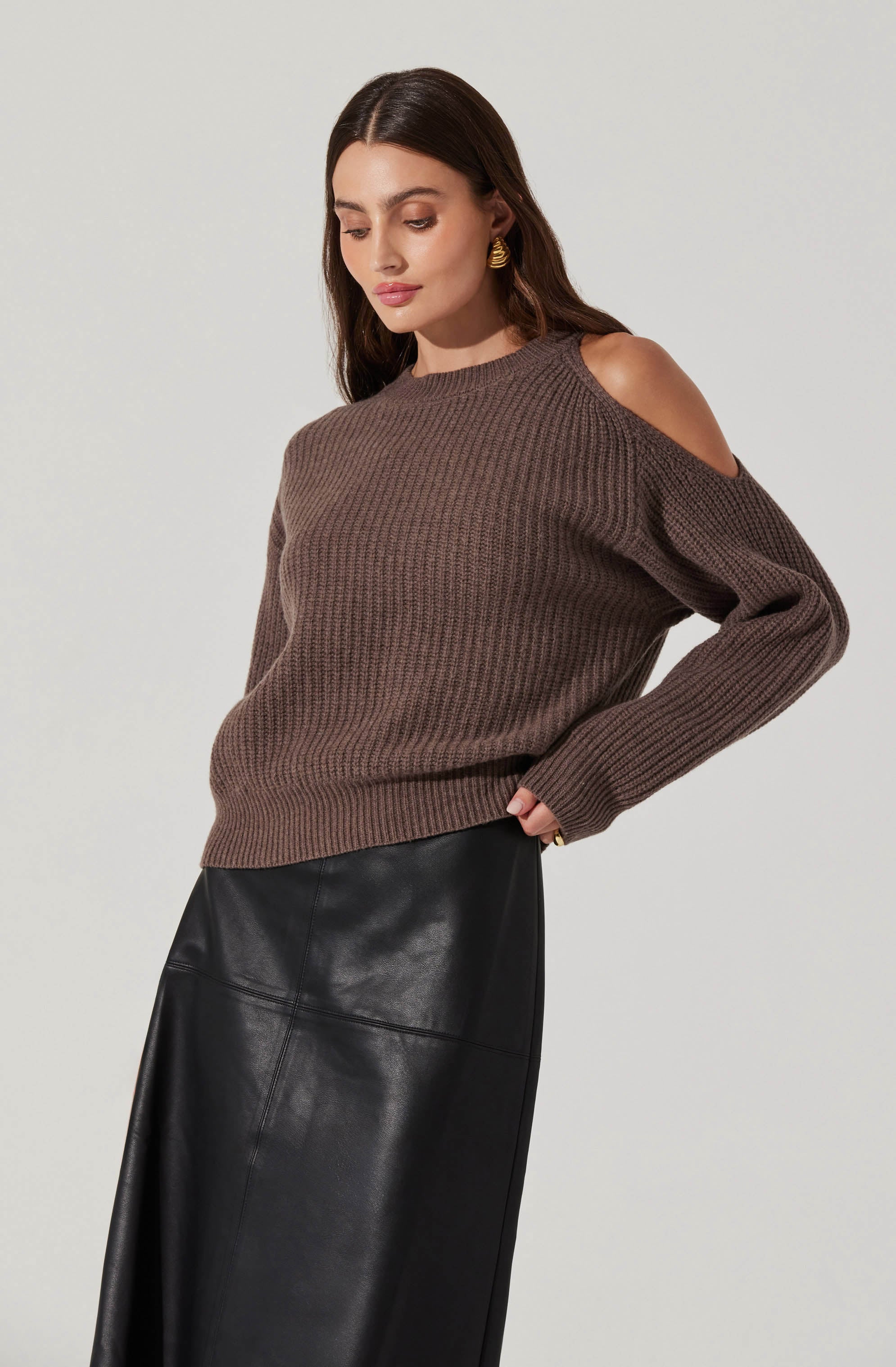 A woman is wearing a brown knit sweater with open shoulder cut-outs and long sleeves. She has paired it with a black leather skirt.