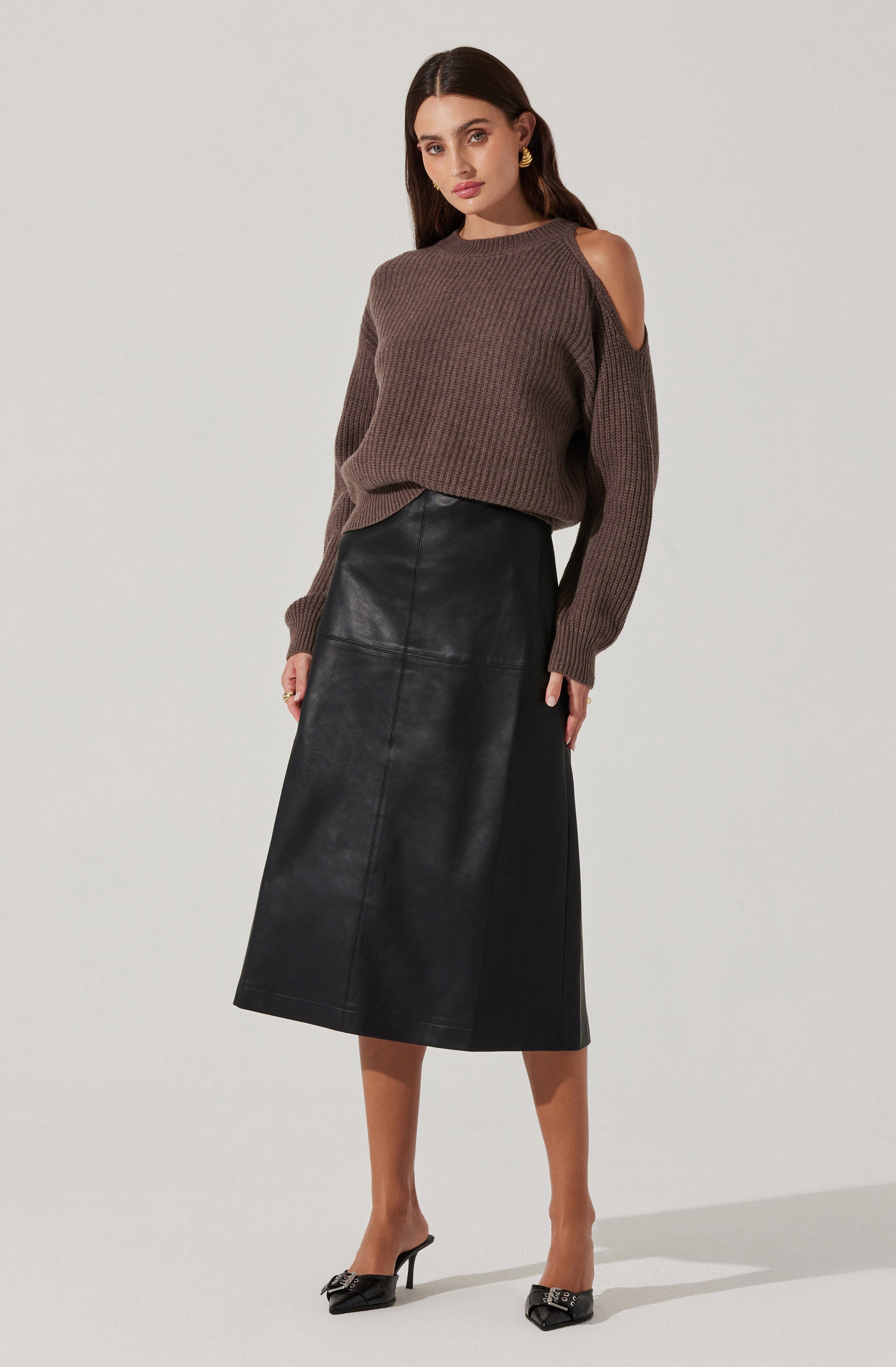 A woman is wearing a brown knit sweater with cut-out shoulder details, paired with a high-waisted black leather midi skirt. She completes the outfit with black pointed-toe heels and gold earrings.