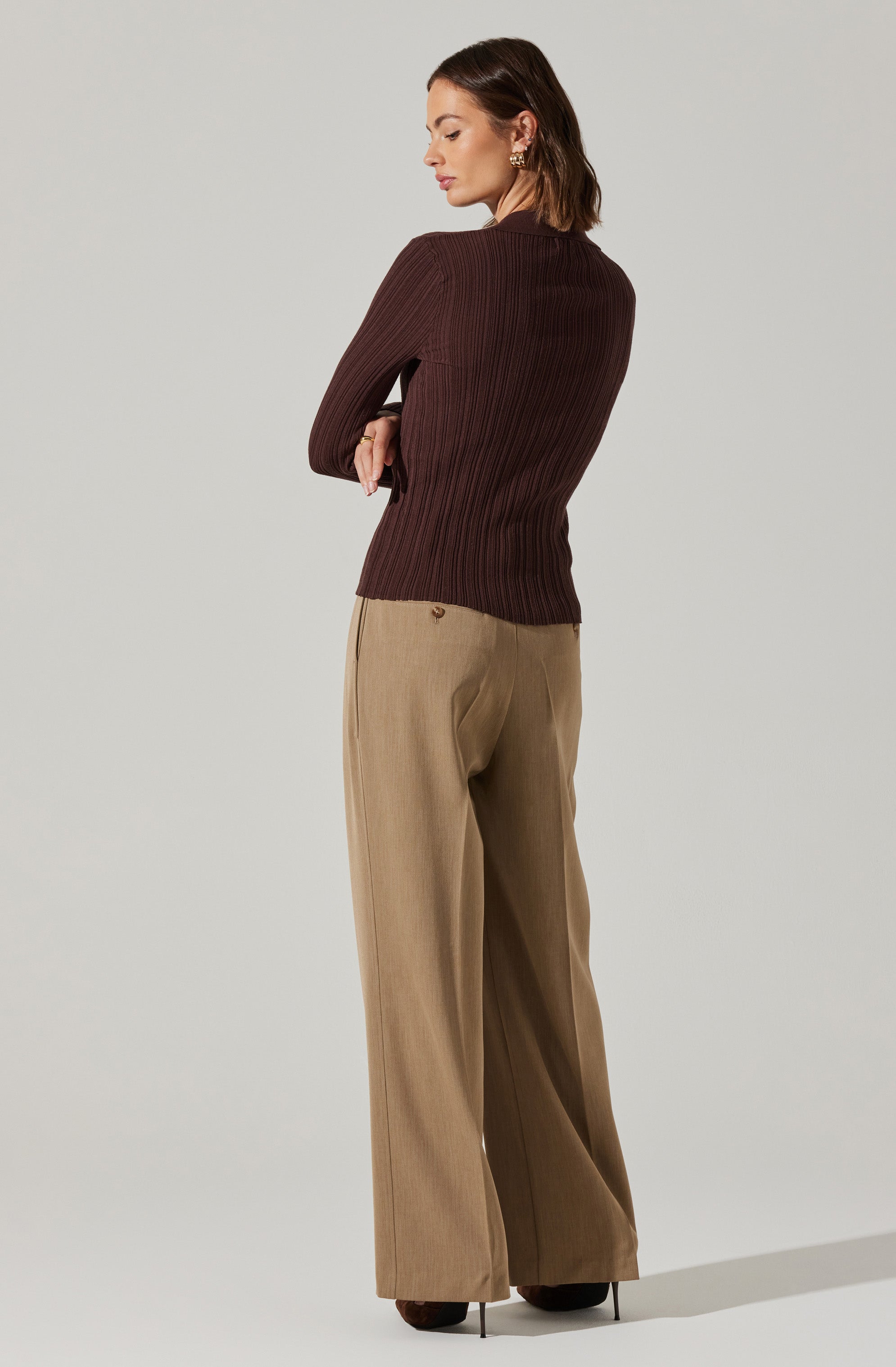A woman is wearing a fitted, long-sleeved, ribbed sweater in a dark brown color and high-waisted, wide-leg beige trousers with a subtle pinstripe pattern. She has earrings and is standing in a neutral pose against a plain light background.