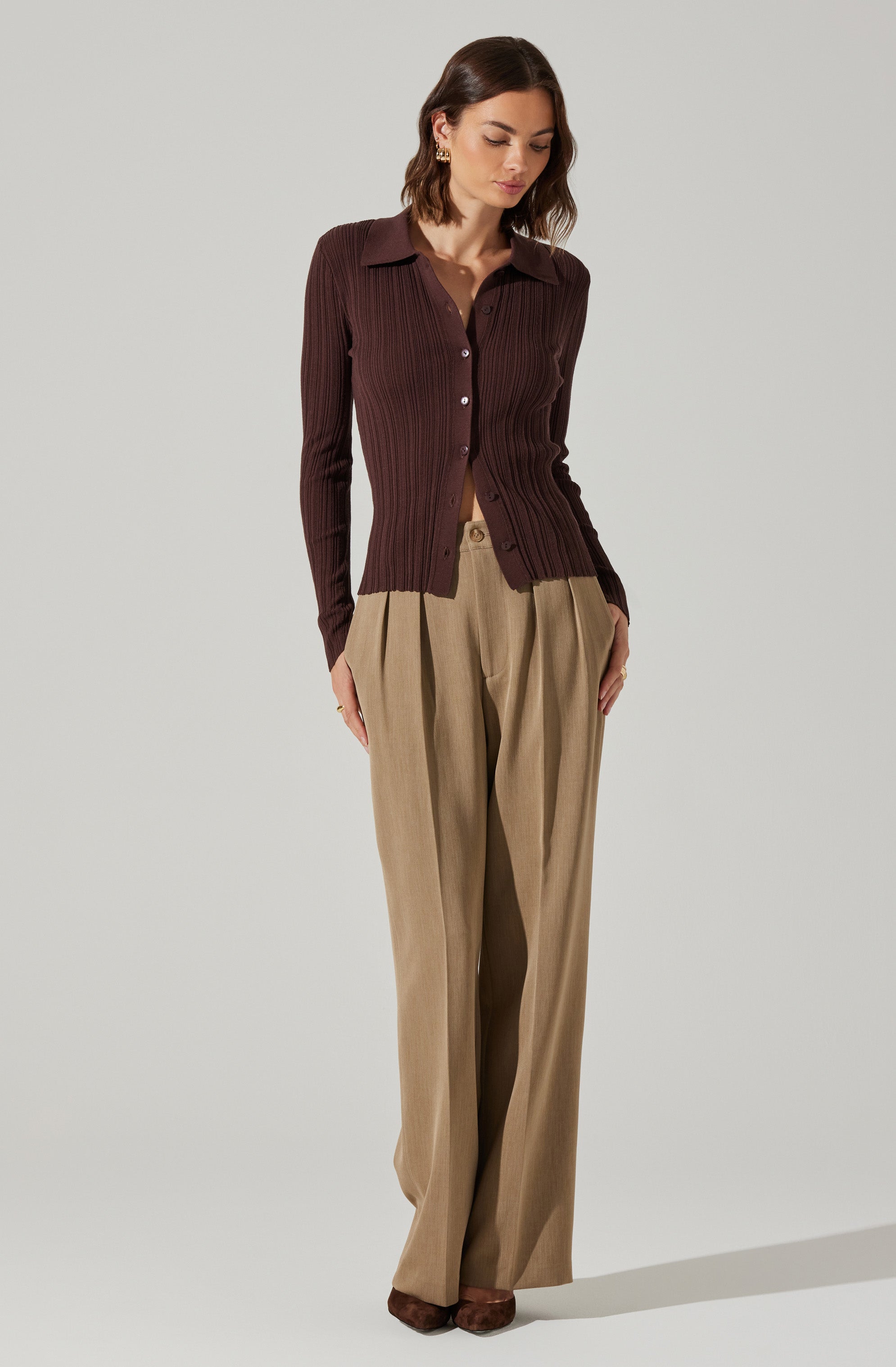 A woman is wearing a fitted, long-sleeved, dark brown ribbed cardigan with a collar and button-down front, paired with high-waisted, wide-leg beige trousers.