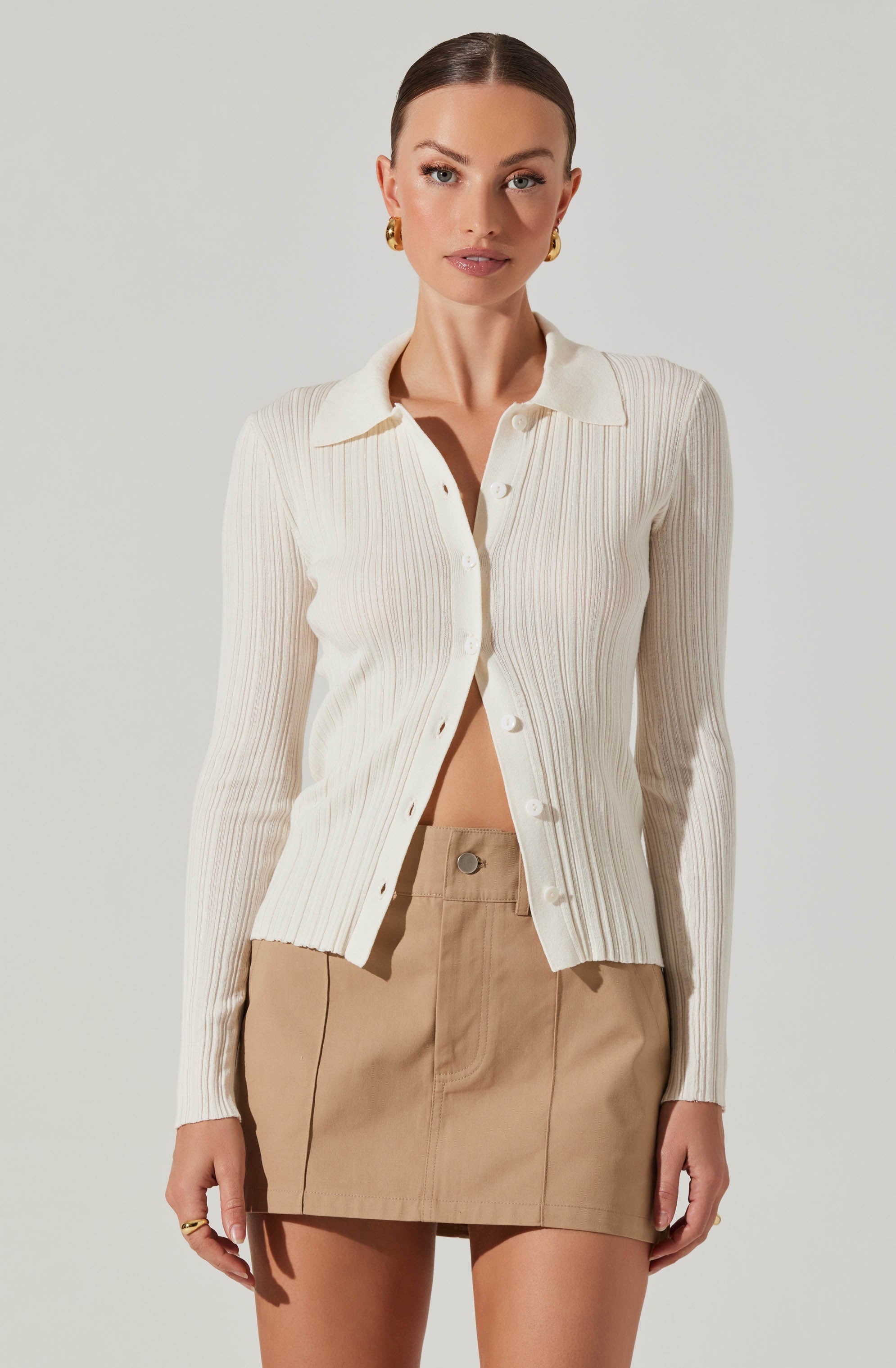 A woman is wearing a cream-colored, ribbed, button-up cardigan with a collar. The cardigan has a fitted style and long sleeves. She pairs it with a high-waisted tan skirt featuring visible stitching details. The overall outfit has a neutral and classic color palette.