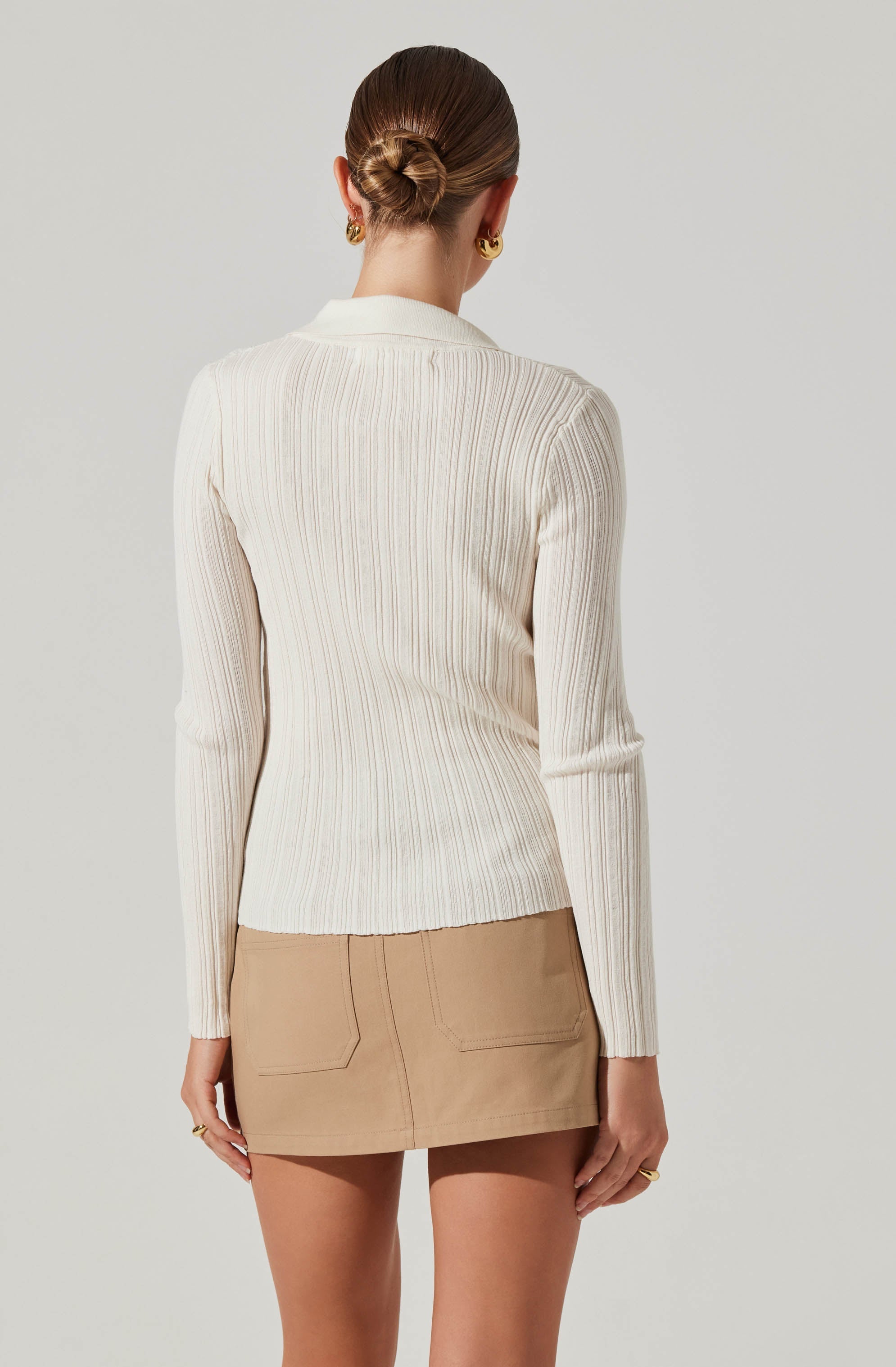 A woman is wearing a long-sleeved, ribbed, cream-colored sweater with vertical stripe patterns. The sweater has a collared neckline. She is also wearing a beige mini skirt with front and back pockets. Her hair is styled in a neat bun, and she is accessorized with gold hoop earrings and rings.