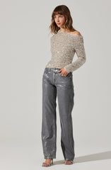A woman is wearing a long-sleeve, off-the-shoulder top that is fully covered in sparkly beads or sequins, giving it a shiny, glamorous appearance. She is also wearing high-waisted, loose-fitting gray jeans with a metallic sheen, paired with open-toe heels.