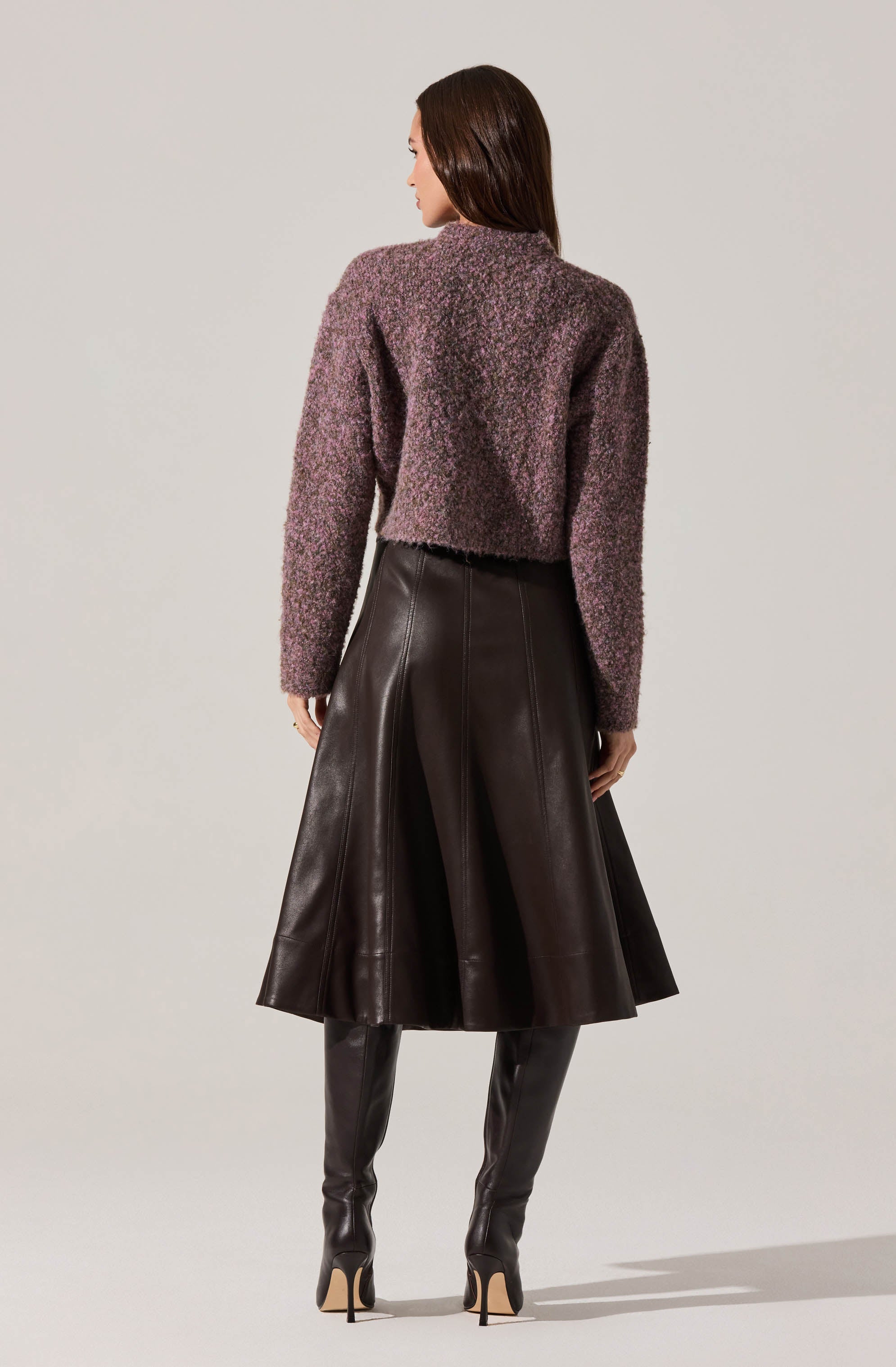 A woman is wearing a textured, long-sleeved, purple and gray knit sweater paired with a high-waisted, midi-length leather skirt that flares out. She completes the outfit with tall, black leather over-the-knee boots with stiletto heels.