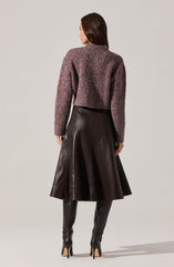 A woman is wearing a textured, long-sleeved, purple and gray knit sweater paired with a high-waisted, midi-length leather skirt that flares out. She completes the outfit with tall, black leather over-the-knee boots with stiletto heels.