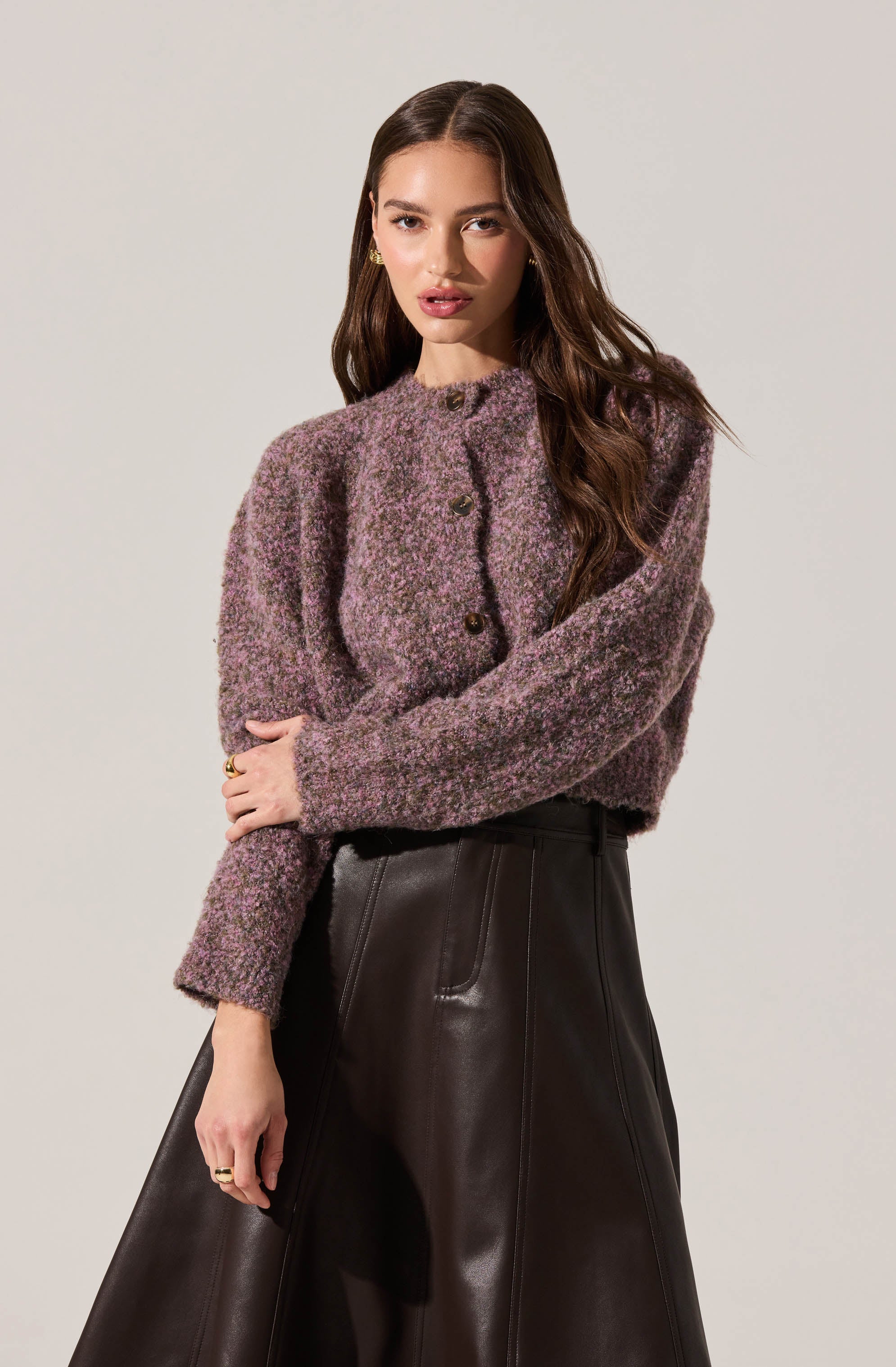 A woman is wearing a textured, long-sleeved cardigan in shades of purple and gray with a high neckline and large buttons, paired with high-waisted, loose-fitting black leather pants.