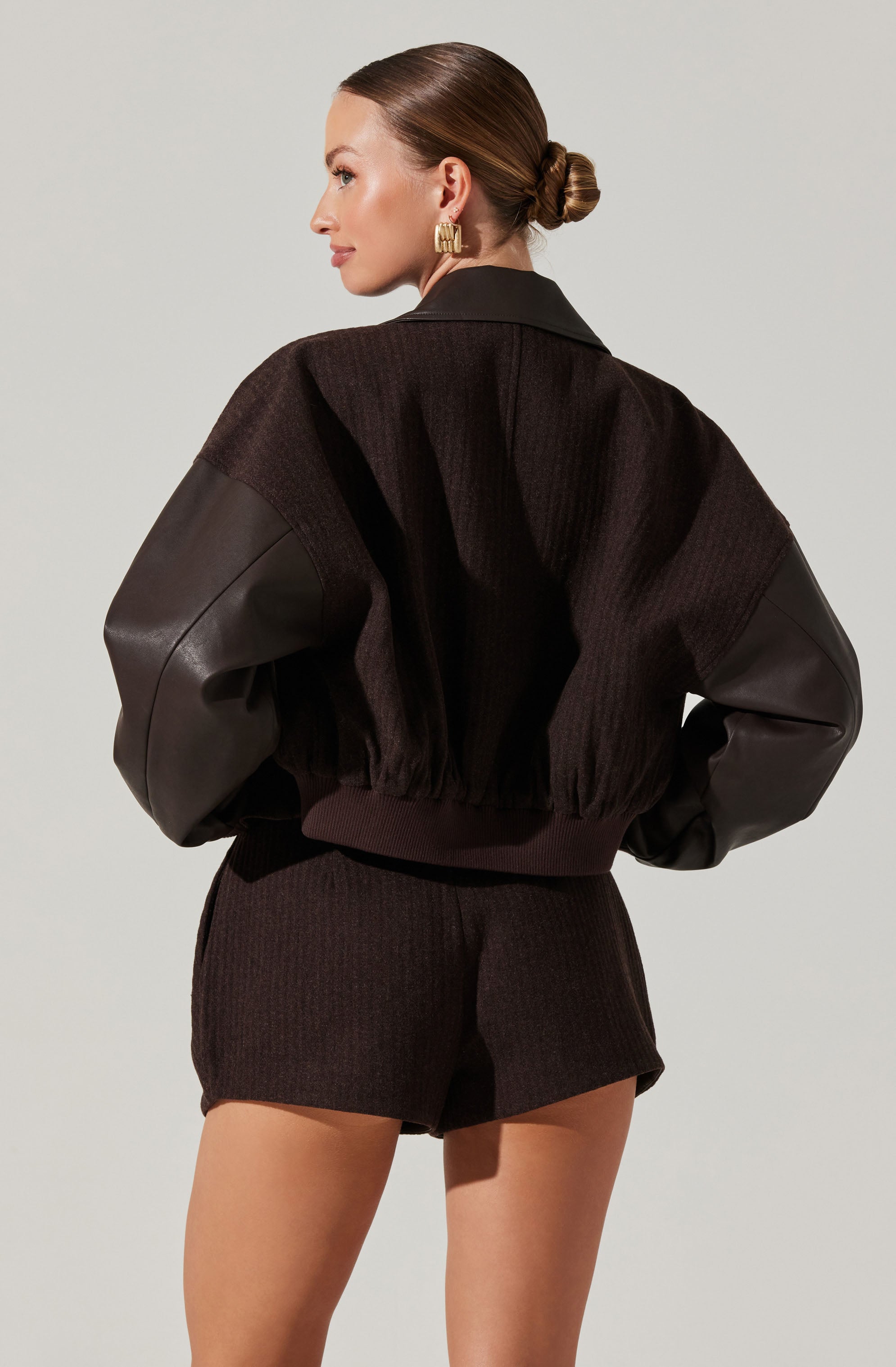 A woman is wearing a black bomber jacket with large, textured sleeves made of leather and knit fabric, featuring a ribbed waistband. She pairs it with matching black knit shorts and has her hair styled in a low, neat bun. She is also wearing statement gold earrings.
