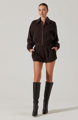 A woman is wearing a dark brown zip-up jacket with a collar, featuring a mix of fabric textures, paired with matching high-waisted shorts that have pleats at the front. She is also wearing tall black leather boots that reach just below her knees.