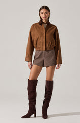 A woman is wearing a brown cropped jacket with a collar and long sleeves. She has on high-waisted shorts with a small checkered pattern. She is also wearing tall, dark brown suede over-the-knee boots with high heels.