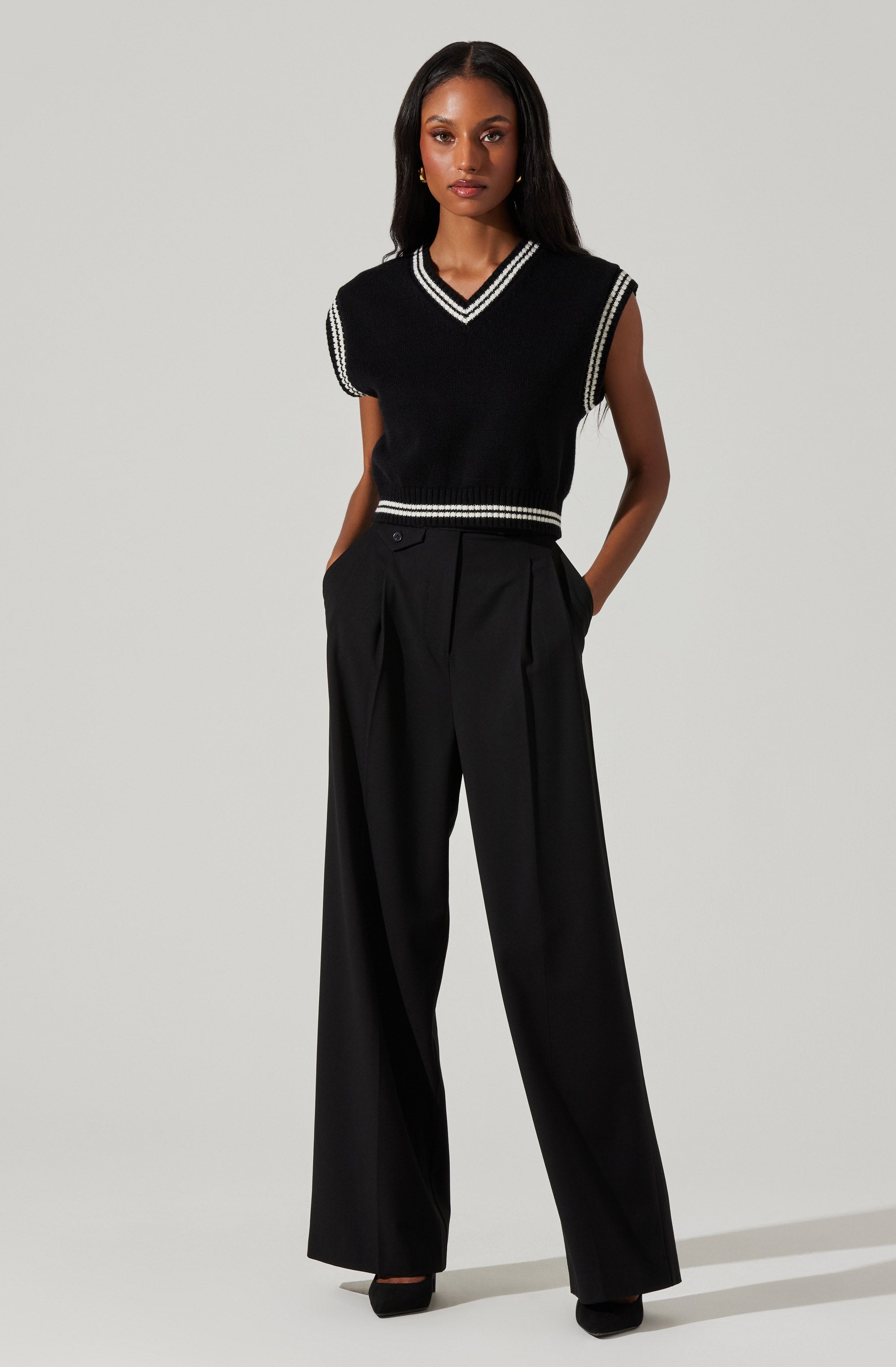 A woman is wearing a sleeveless black knit vest with white stripes along the V-neckline, armholes, and hem. She pairs it with high-waisted, wide-leg black trousers.