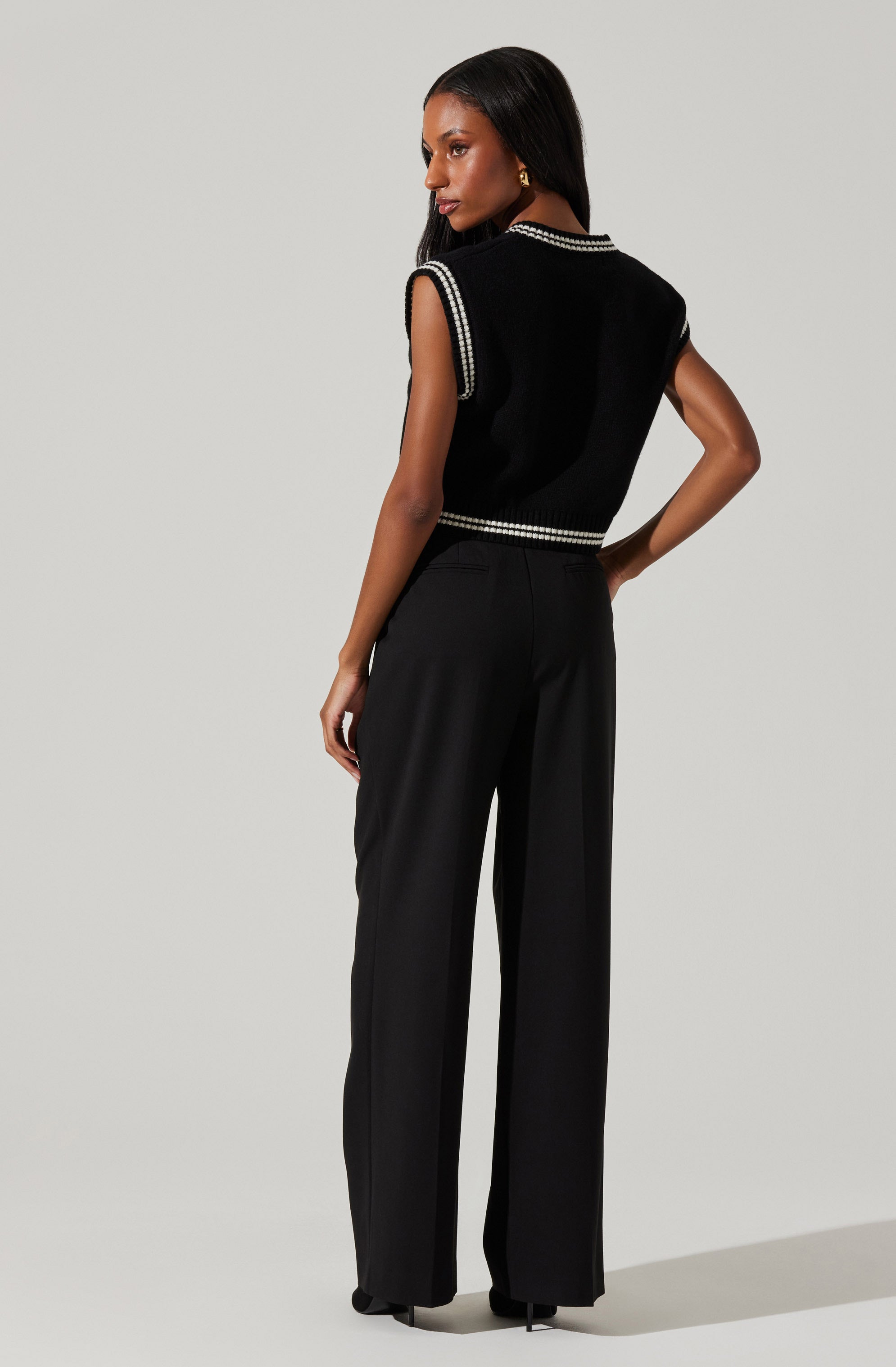 A woman is wearing a sleeveless black top with a round neckline, accented with white trim along the edges. She pairs the top with high-waisted black wide-leg trousers.
