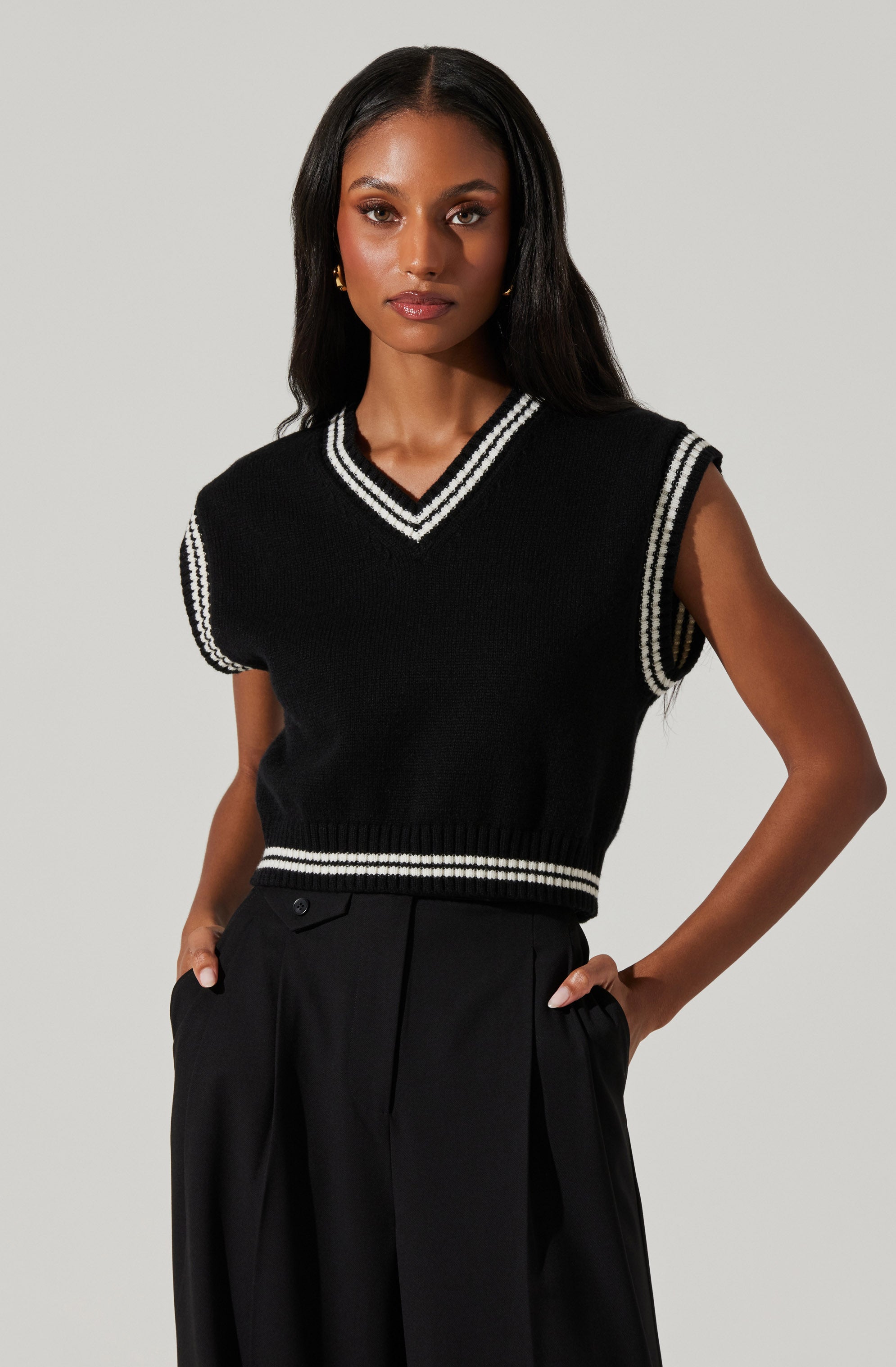 A woman is wearing a black sleeveless sweater vest with white and black striped trim around the V-neckline, armholes, and hem. She pairs it with black high-waisted trousers.