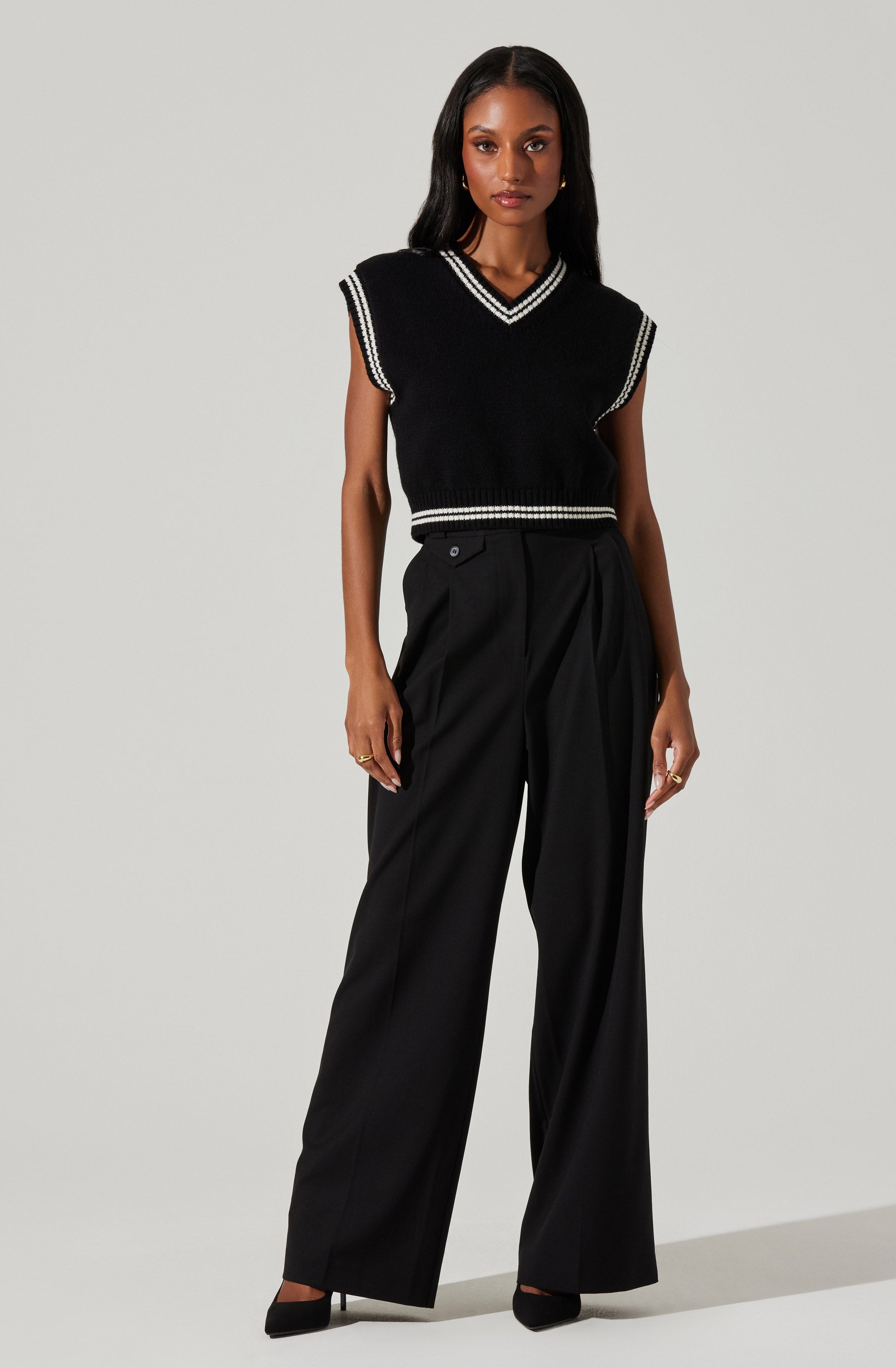 A woman is wearing a sleeveless black vest with white trim along the neckline, armholes, and hem. She pairs the vest with high-waisted, wide-leg black trousers. The outfit is accessorized with gold jewelry and black pointed-toe shoes.