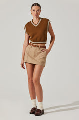 A woman is wearing a sleeveless, brown, knitted vest with white and beige stripes along the V-neckline, armholes, and hem. She pairs this with beige high-waisted shorts with front pleats and a belt, and completes the look with beige socks and dark brown loafers.