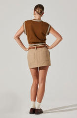 A person is wearing a sleeveless, brown knitted vest with white and beige stripe details along the armholes and neckline, paired with a beige skirt that has large patch pockets on the back. They are also wearing cream-colored socks and dark brown shoes.
