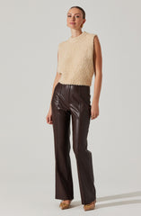 A woman is wearing a sleeveless, high-neck, fuzzy beige top paired with high-waisted, dark brown leather pants that have a straight-leg fit. She is accessorized with gold earrings and a ring, and is wearing pointed-toe heels with a metallic detail.