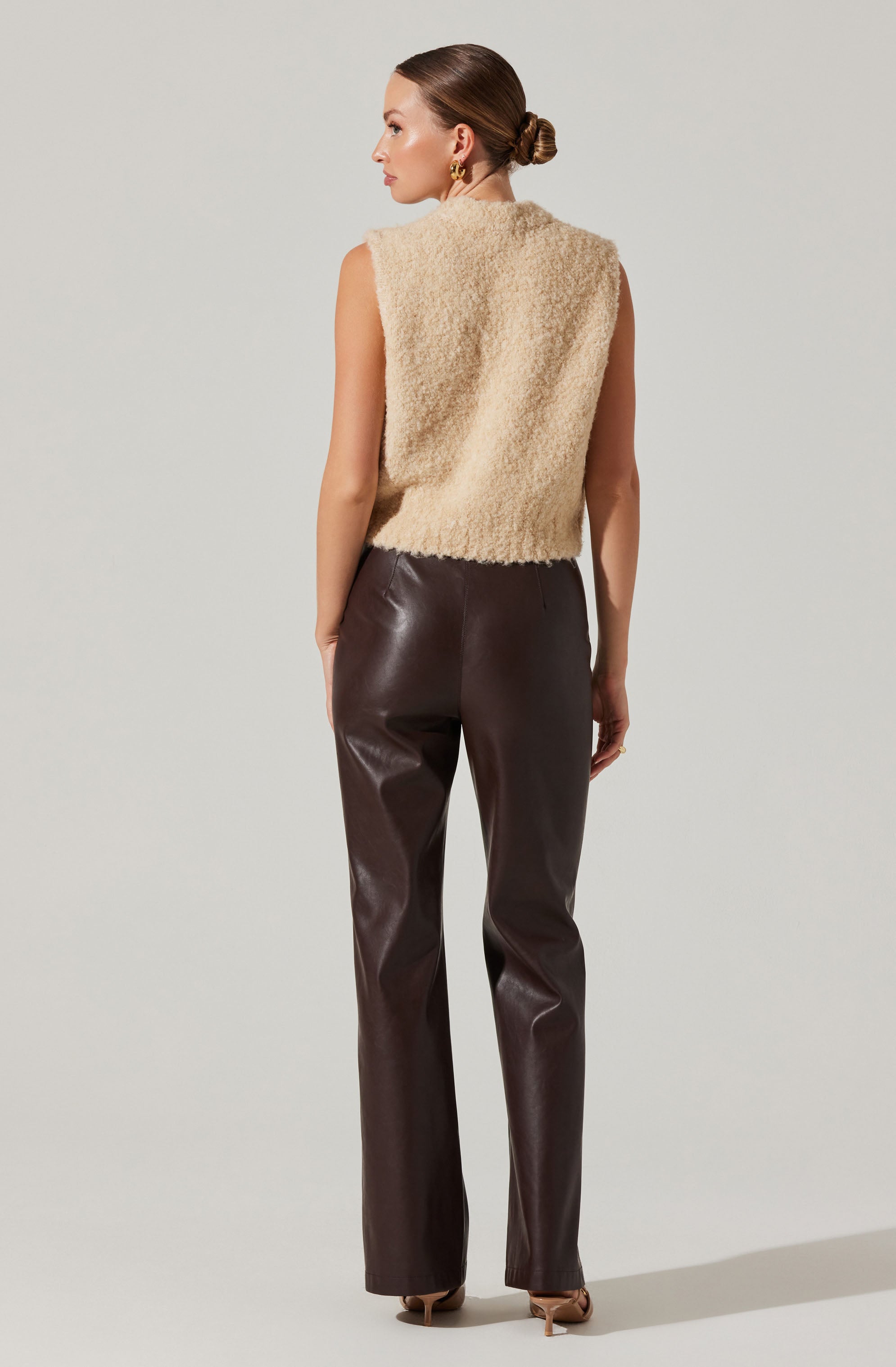A woman is wearing a sleeveless, beige, fuzzy vest made of a soft, textured material. She pairs it with high-waisted, dark brown leather pants that fit snugly through the hips and thighs, and have a straight-leg cut. She is also wearing beige high-heeled shoes. Her hair is styled in a neat low bun, and she has gold hoop earrings.