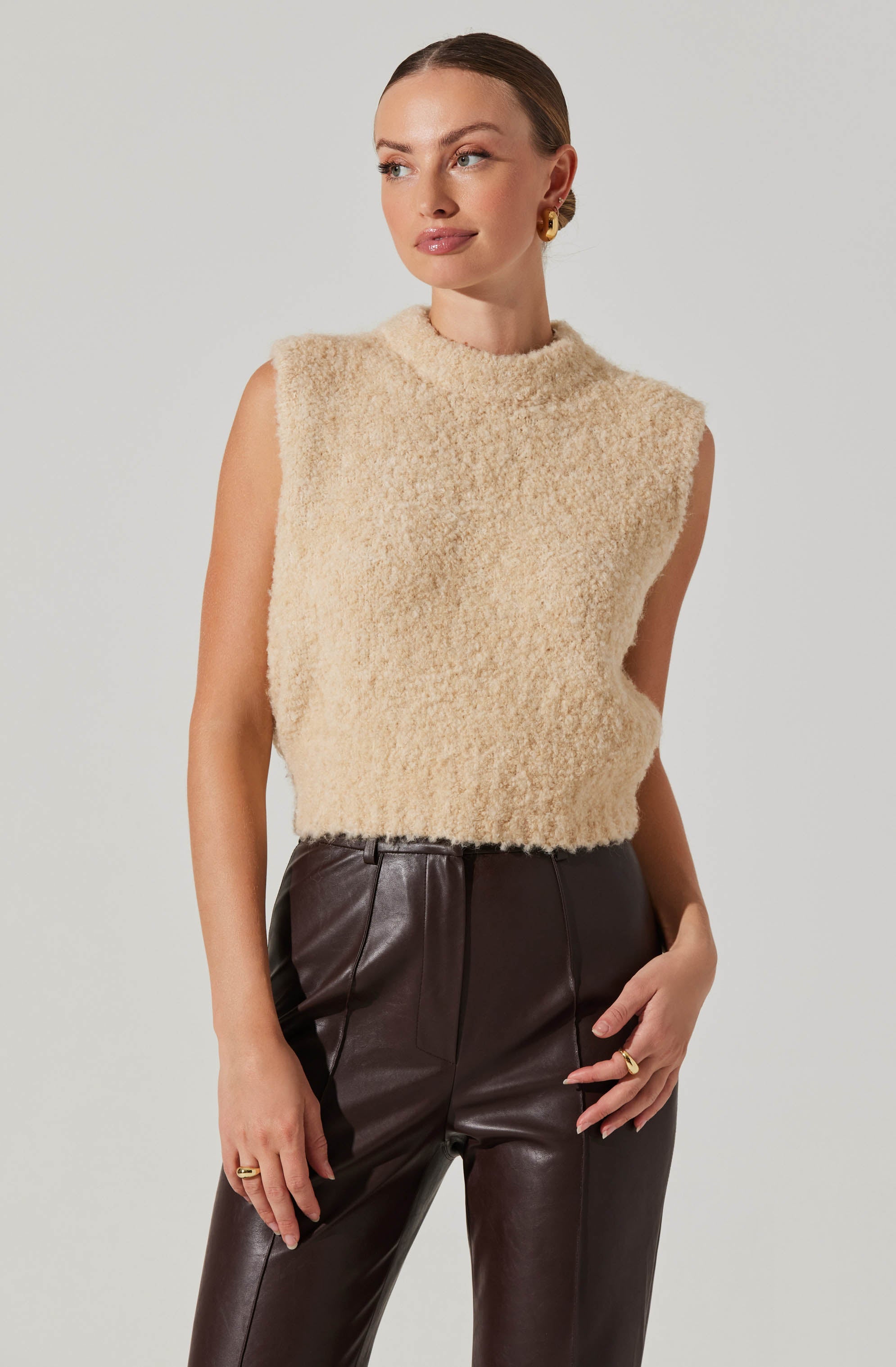 A woman is wearing a sleeveless, high-neck, cream-colored, fuzzy sweater paired with high-waisted, dark brown leather pants.