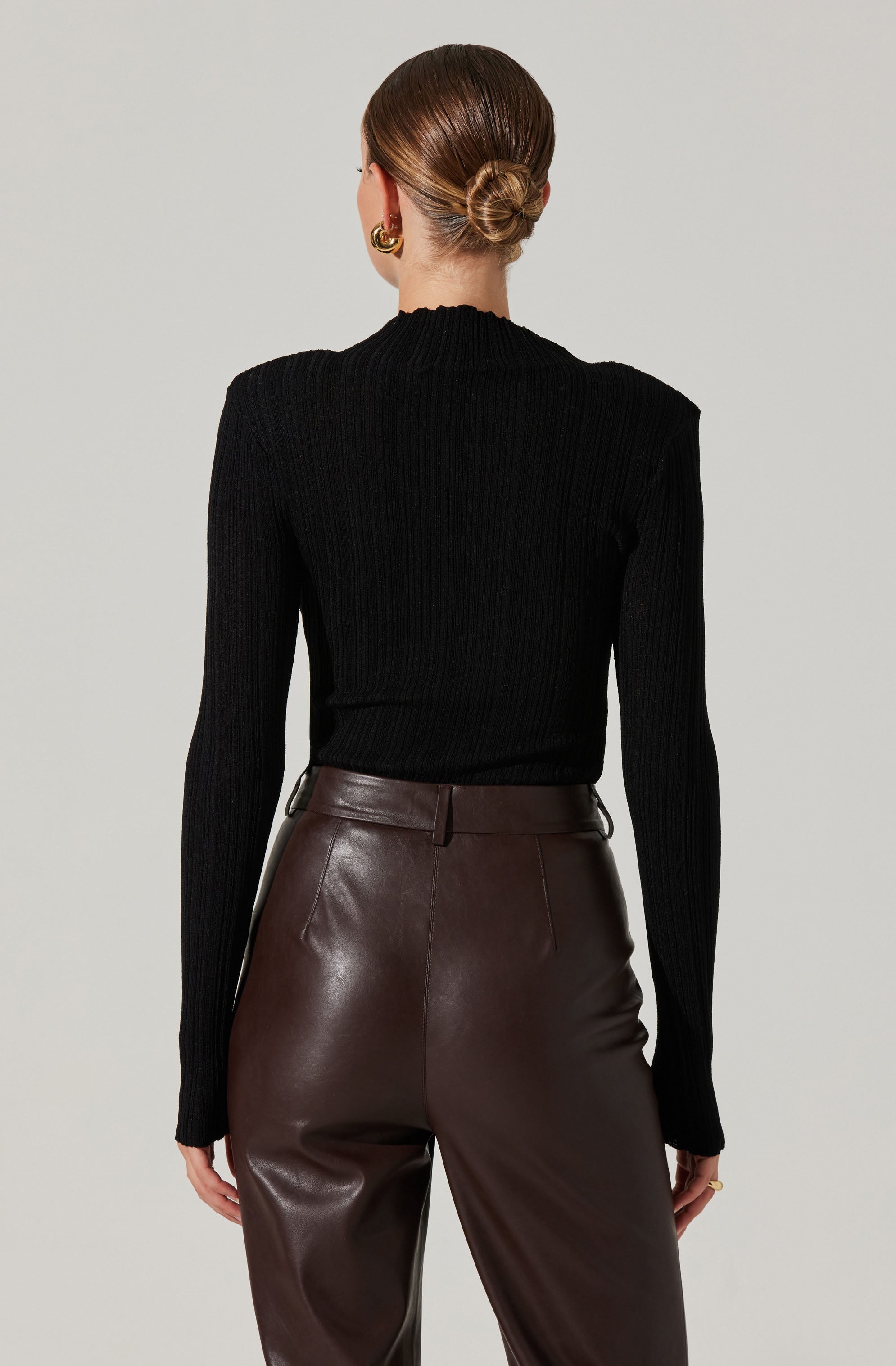 A woman is wearing a fitted black ribbed sweater with a high neckline and long sleeves. She pairs it with high-waisted, dark brown leather pants that have a smooth finish and a waistband with belt loops. Her hair is styled in a low, neat bun, and she is accessorized with large, gold hoop earrings.