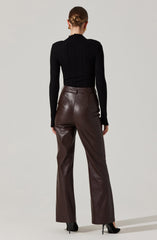A person is wearing a fitted, long-sleeve black ribbed turtleneck sweater paired with high-waisted, wide-leg brown leather pants. They are also wearing black pointed high-heeled shoes and gold earrings. Their hair is styled in a neat bun.