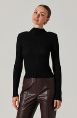 A woman is wearing a fitted black ribbed turtleneck sweater with long sleeves and a high neckline. She is also wearing high-waisted brown leather pants with visible vertical seams.