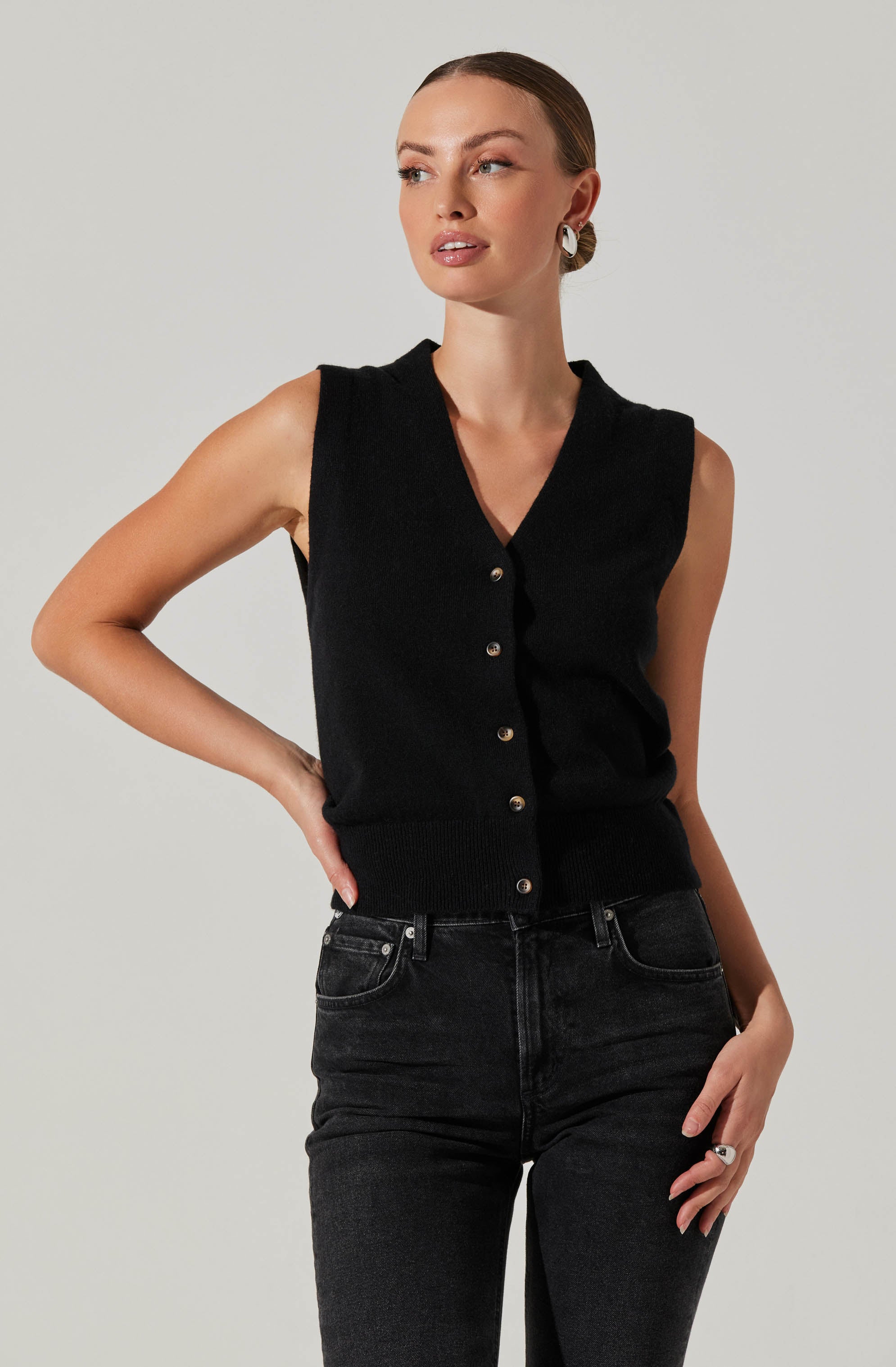A woman is wearing a sleeveless black knit vest with a V-neckline and a row of buttons down the front. She is paired it with high-waisted black jeans.
