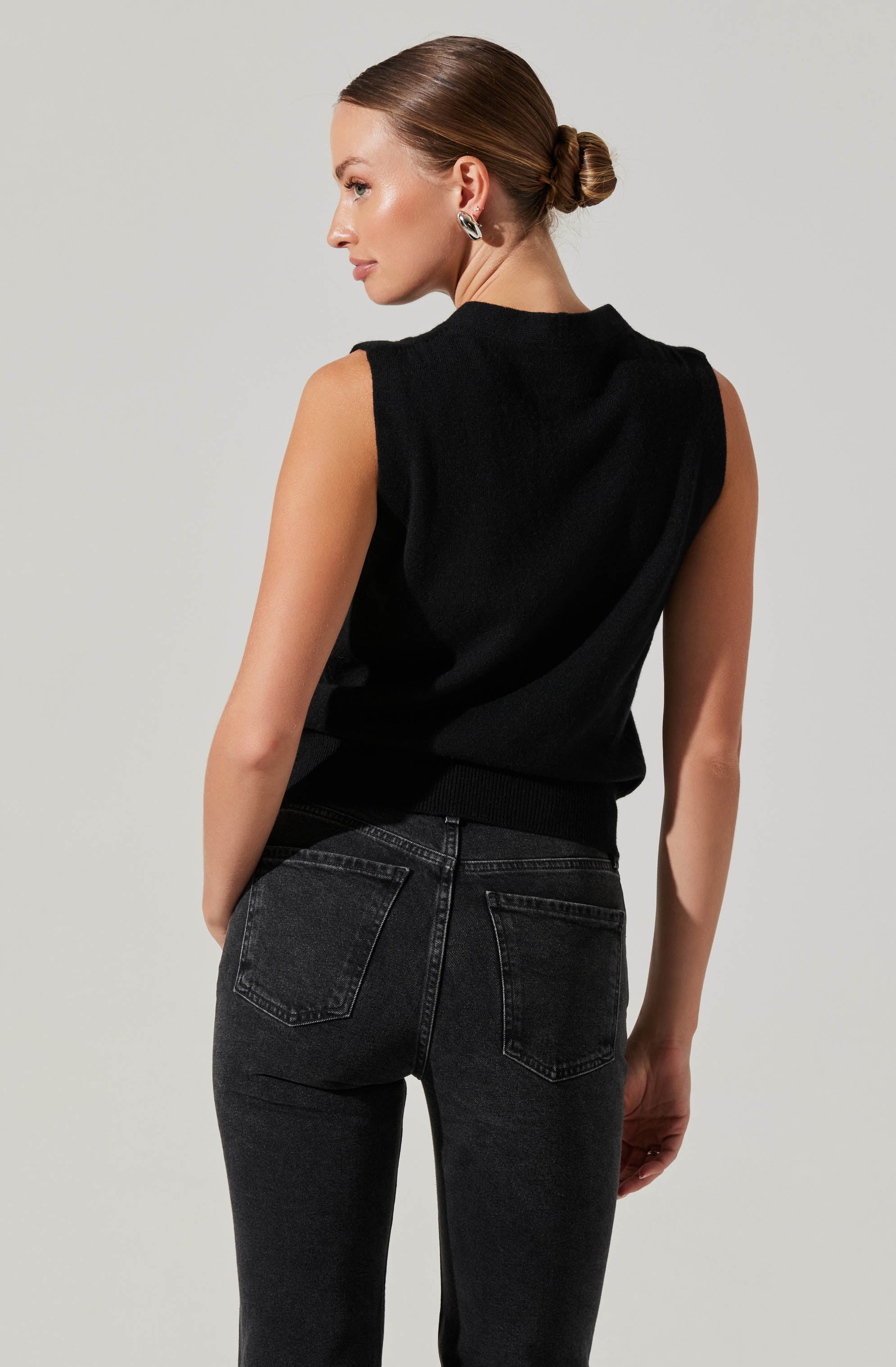 A woman is wearing a sleeveless black top with a high neckline and a ribbed hem, paired with high-waisted, faded black jeans. She has her hair tied back in a sleek bun and is accessorized with hoop earrings.