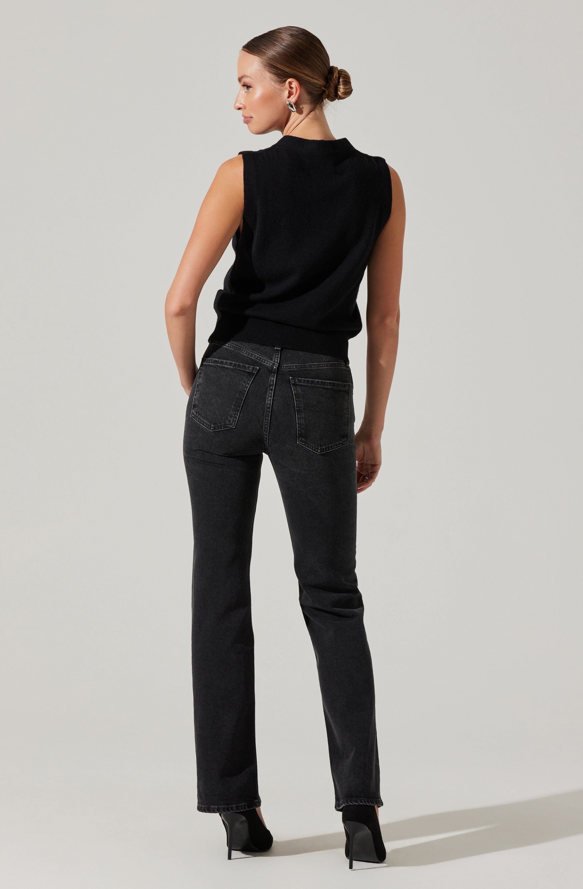 A woman is wearing a sleeveless black top with a high neckline and a slightly loose fit. She pairs this with dark wash high-waisted jeans that have a straight-leg cut and prominent back pockets. She is also wearing black high-heeled shoes. Her hair is styled in a neat bun, and she has on large, possibly metallic, earrings.