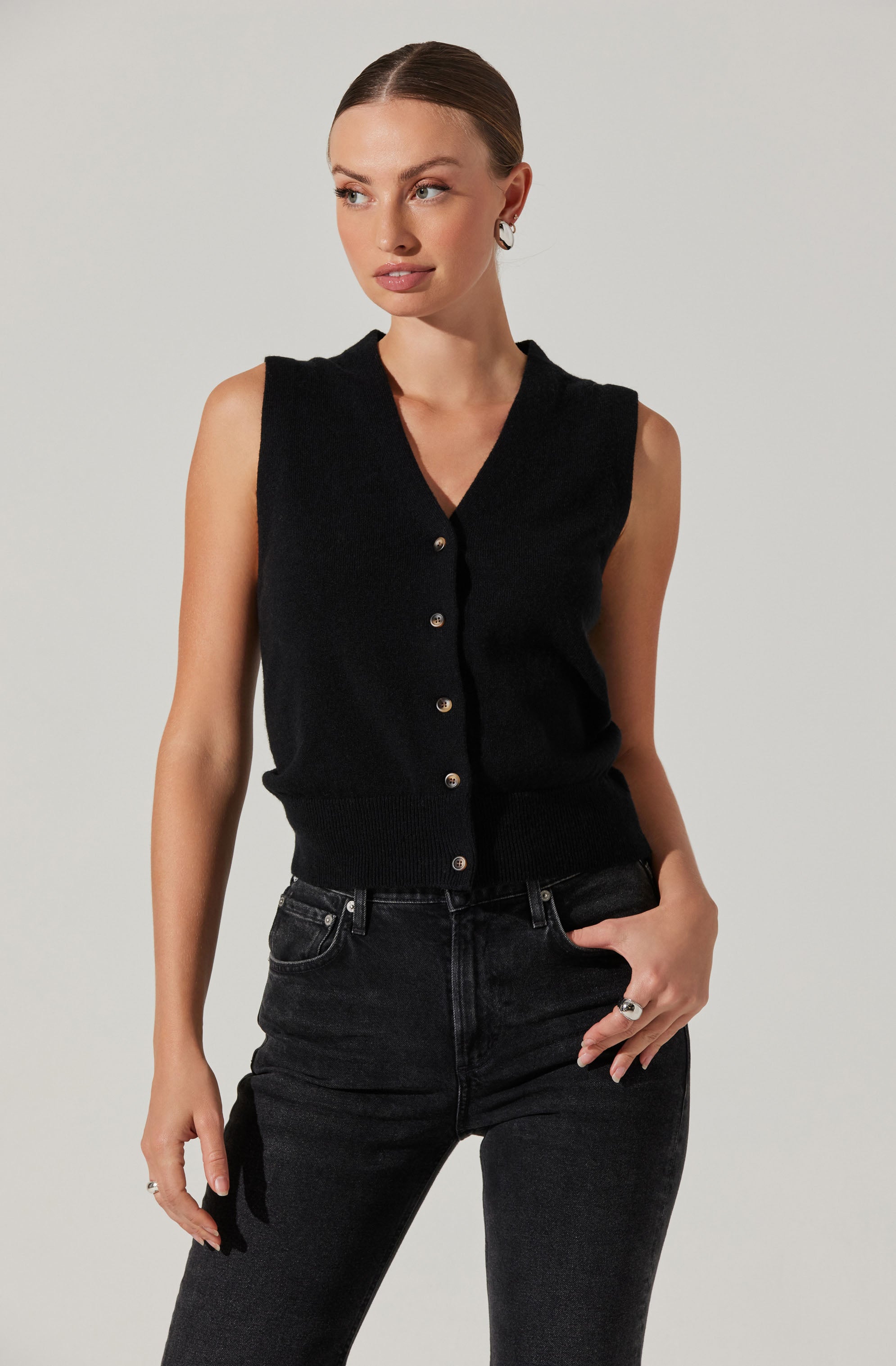 A woman is wearing a sleeveless black knitted vest with a V-neckline and button closure down the front. She pairs it with high-waisted black denim jeans.