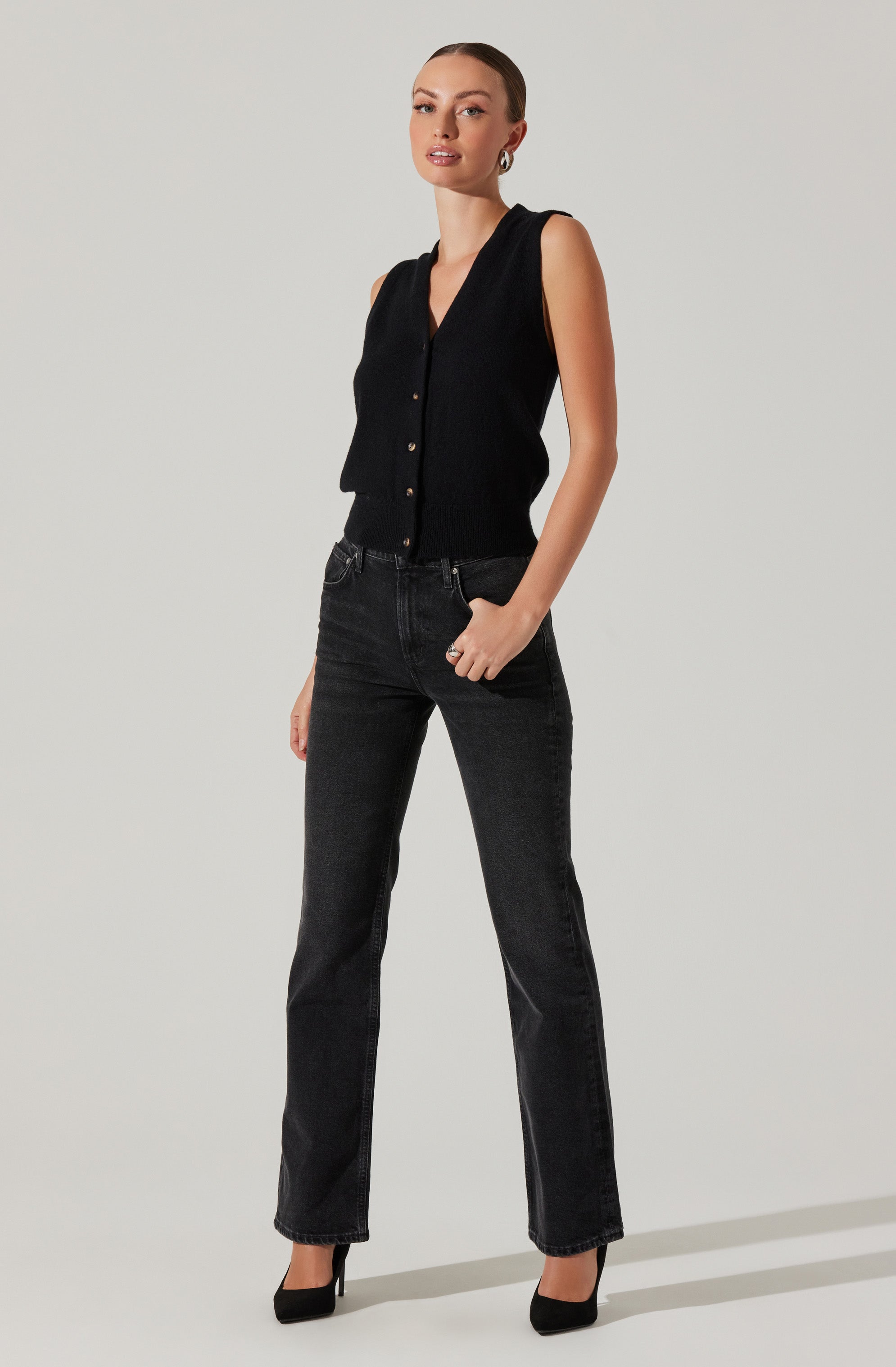 A woman is wearing a sleeveless black button-up vest with a V-neckline, paired with high-waisted black jeans and black high heels. She accessorizes with hoop earrings and a ring on her right hand.