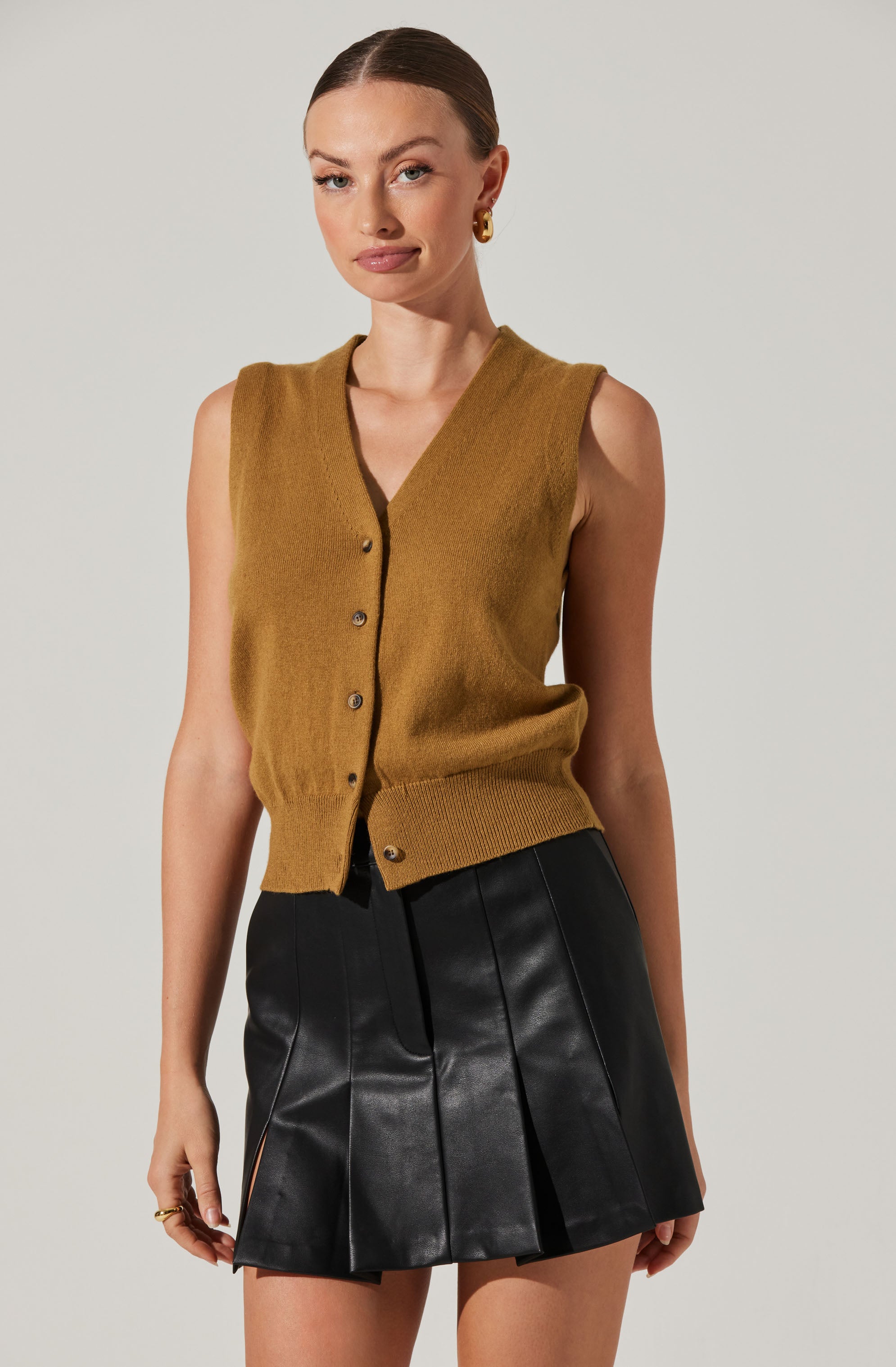A woman is wearing a sleeveless, mustard-colored V-neck cardigan with a button-up front and a ribbed waistband, paired with a black pleated leather skirt.