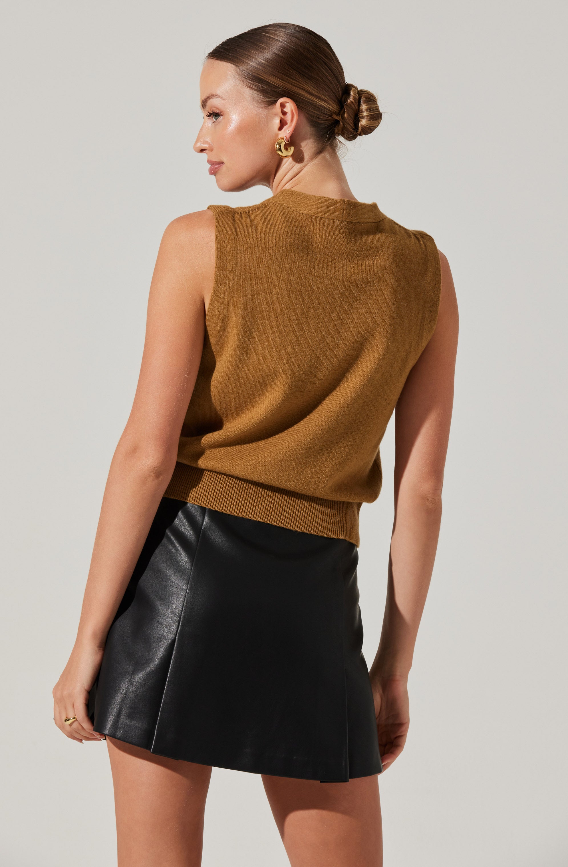 A woman is wearing a sleeveless, brown knitted top with a ribbed hem, paired with a high-waisted black leather mini skirt.