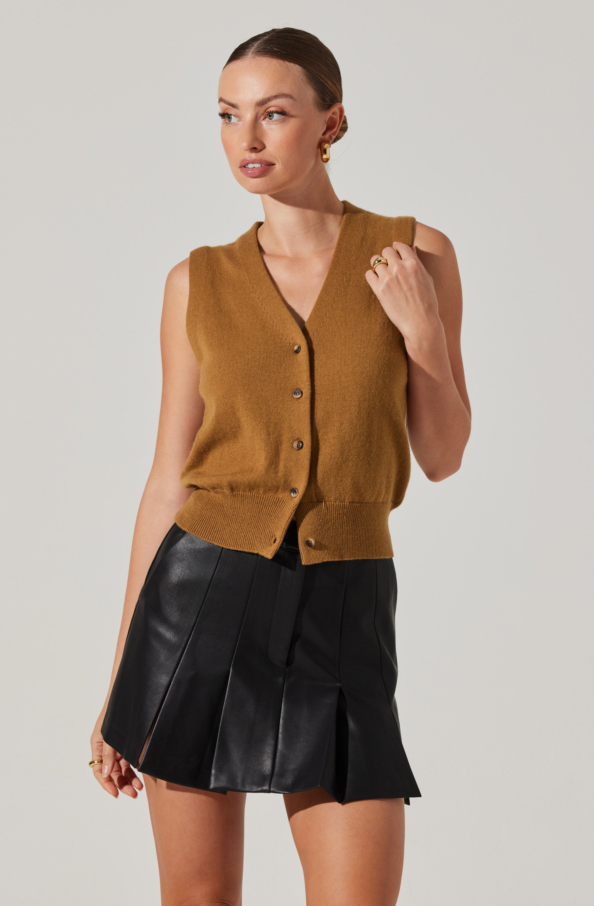 A woman is wearing a sleeveless, button-up vest in a warm brown color, with a V-neckline and ribbed hem. She is also wearing black leather shorts with pleats and a drawstring waistband.