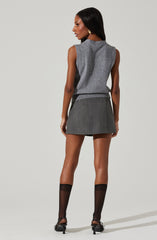 A woman is wearing a sleeveless gray top with a high neckline and a loose fit. She pairs it with a dark gray mini skirt that has a subtle plaid pattern. She is also wearing sheer black knee-high socks and black high-heeled shoes. Her hair is long and straight, and she has large gold hoop earrings.