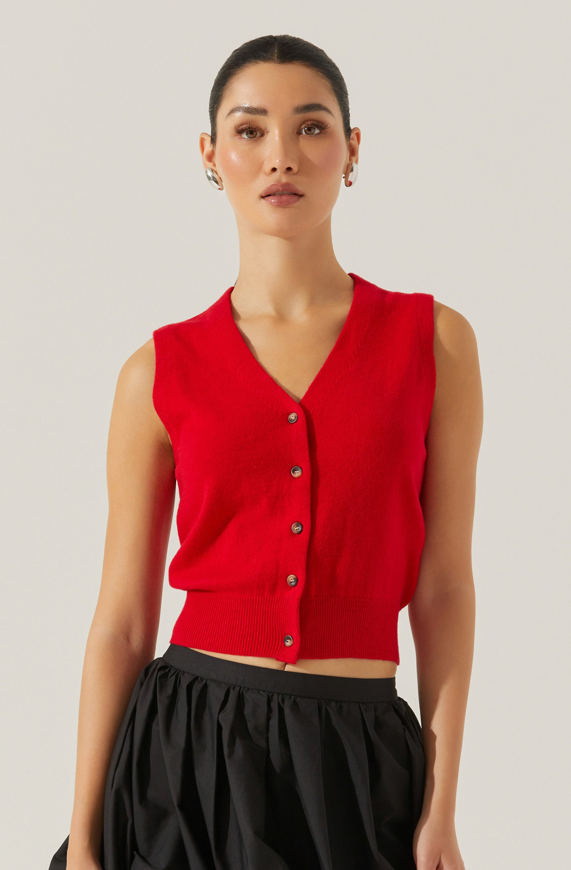 A woman is wearing a sleeveless red knit vest with a V-neckline and button-down front. The vest has a fitted ribbed hem at the waist, creating a cropped look. She is paired with black, loose, gathered pants.