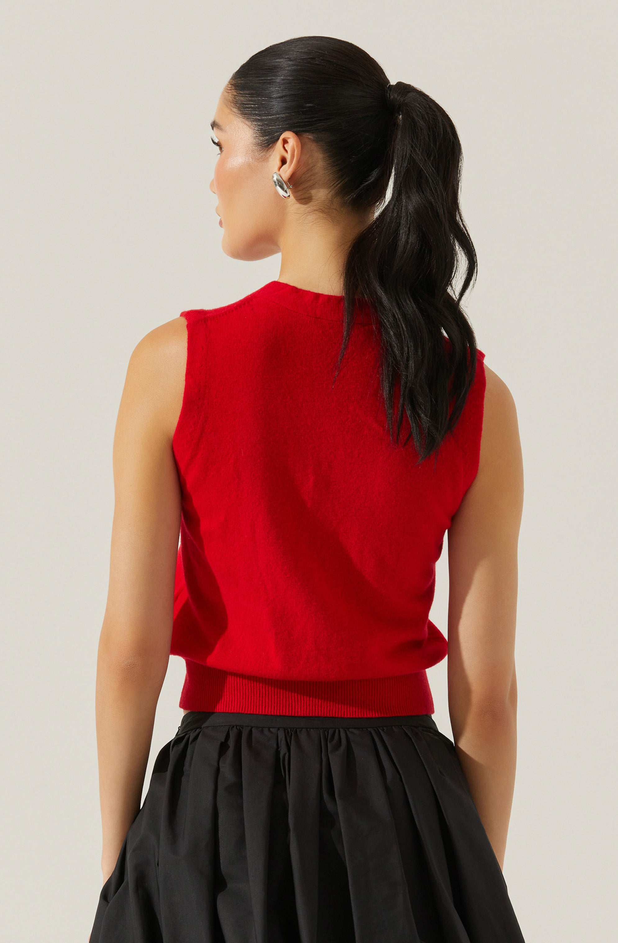 A woman with dark, wavy hair tied in a high ponytail is wearing a sleeveless, bright red knit sweater with a ribbed hem and armholes. She is also wearing a black, pieced, gathered skirt and silver hoop earrings.