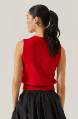 A woman with dark, wavy hair tied in a high ponytail is wearing a sleeveless, bright red knit sweater with a ribbed hem and armholes. She is also wearing a black, pieced, gathered skirt and silver hoop earrings.