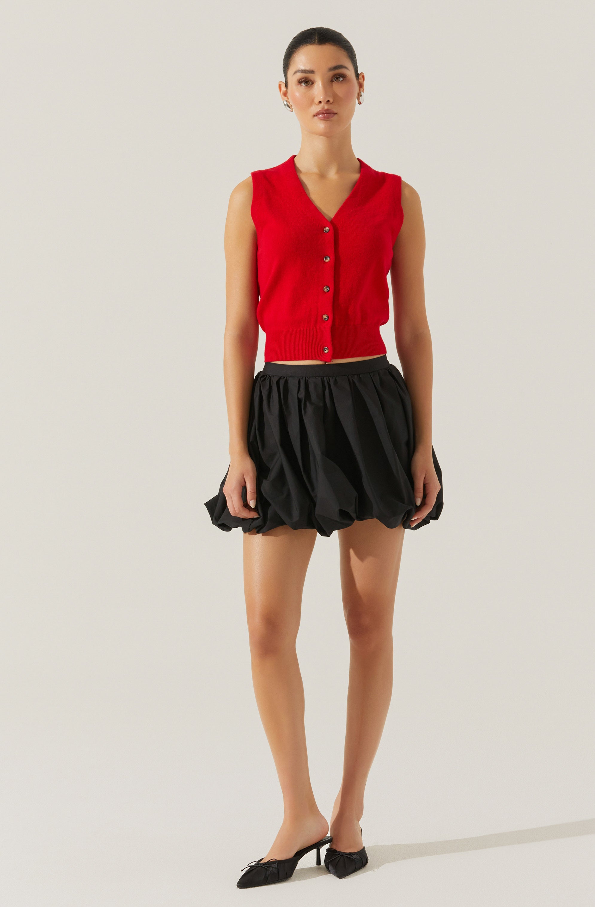 A woman is wearing a sleeveless, red button-up vest with a V-neckline. She pairs it with a high-waisted, black, puffed skirt that has a gathered, voluminous design. She completes her outfit with black pointed-toe heels featuring a small bow detail.