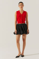 A woman is wearing a sleeveless, red button-up vest with a V-neckline. She pairs it with a high-waisted, black, puffed skirt that has a gathered, voluminous design. She completes her outfit with black pointed-toe heels featuring a small bow detail.