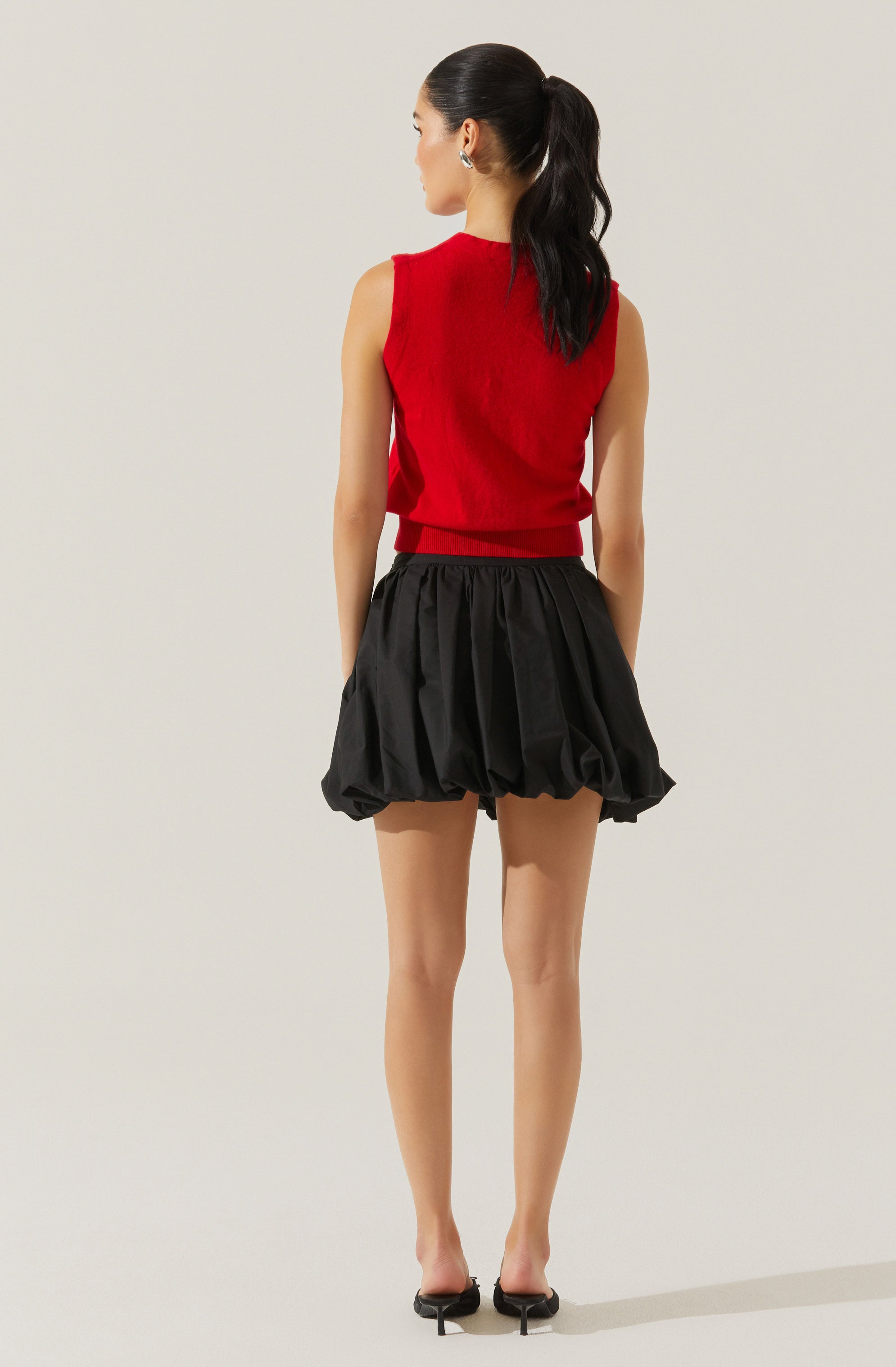 A woman is wearing a sleeveless, red knit top with a ribbed hem, paired with black, voluminous, bubble shorts that have an elastic waistband and a gathered, puffed appearance. She has her hair tied back in a high ponytail and is wearing black high heels.
