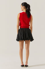 A woman is wearing a sleeveless, red knit top with a ribbed hem, paired with black, voluminous, bubble shorts that have an elastic waistband and a gathered, puffed appearance. She has her hair tied back in a high ponytail and is wearing black high heels.