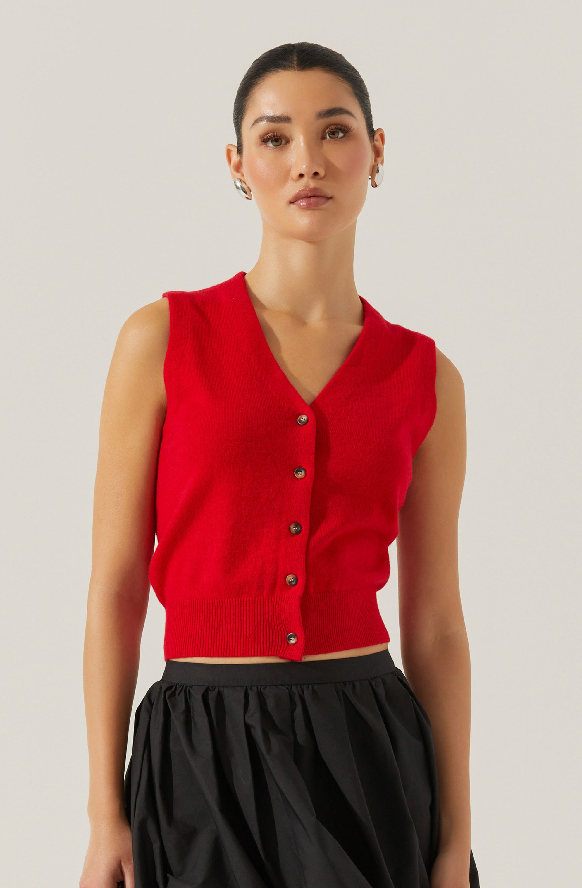 A woman is wearing a sleeveless red knit vest with a V-neckline and a row of metallic buttons down the front. The vest is fitted at the waist, ending just above the waistline. She pairs it with high-waisted, loose-fitting black pants that have a gathered, slightly puffy appearance.