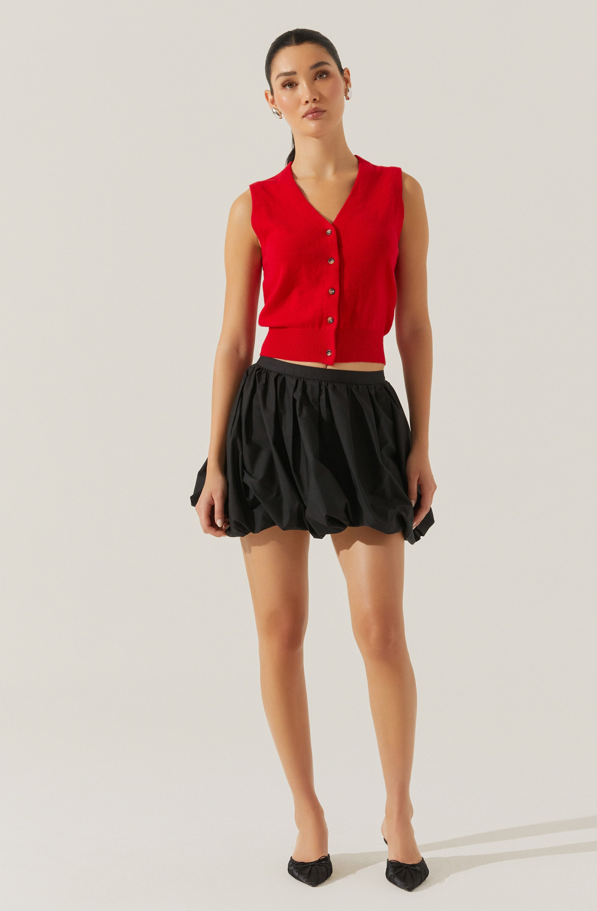 A woman is wearing a sleeveless, red V-neck button-up sweater vest paired with a black, voluminous, ruched mini skirt. She is also wearing black pointed-toe heels with small bow accents.