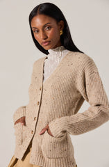 A woman is wearing a beige, chunky knit cardigan with a speckled pattern, featuring large front pockets and wooden buttons down the front. Underneath, she has on a high-neck, textured white blouse. She also has gold earrings and her hair is long and straight.