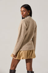 A woman is wearing a beige, chunky knit sweater with a high neckline and long sleeves. She has paired it with a beige, puffy, mini skirt that has a gathered, voluminous appearance. She is also wearing large, gold square earrings.