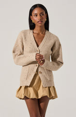 A woman is wearing a beige, chunky knit cardigan with a V-neckline, buttoned in the front, featuring a speckled pattern and ribbed cuffs. She pairs it with a tan-colored, puffy, bubble-hem skirt made of lightweight fabric. She is also accessorized with gold earrings and rings.